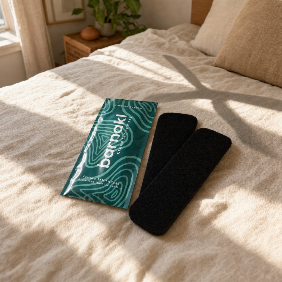 A teal Barnakl pack and two jet-black carbon pads staged on a sunlit bed with a ceiling fan in frame