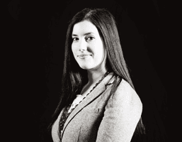 A black and white portrait of a woman with long dark hair, wearing a light-colored blazer and a patterned necklace. She is looking slightly to her left. The background is uniformly dark.