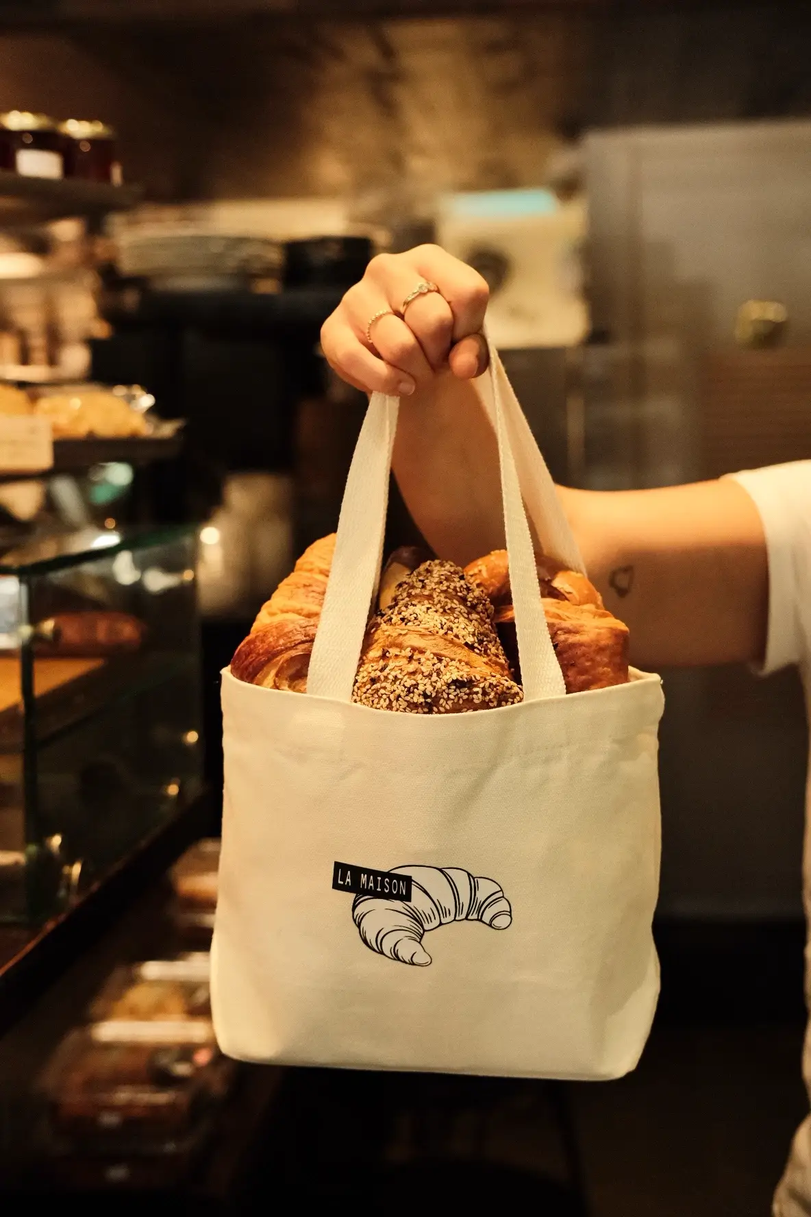 צילום לייף-סטייל של יד אוחזת בתיק בד מלא במאפים וקרואסונים טריים בתוך מאפייה, צילום אווירה ומזון מקצועי - Shiriz שירי עציץ