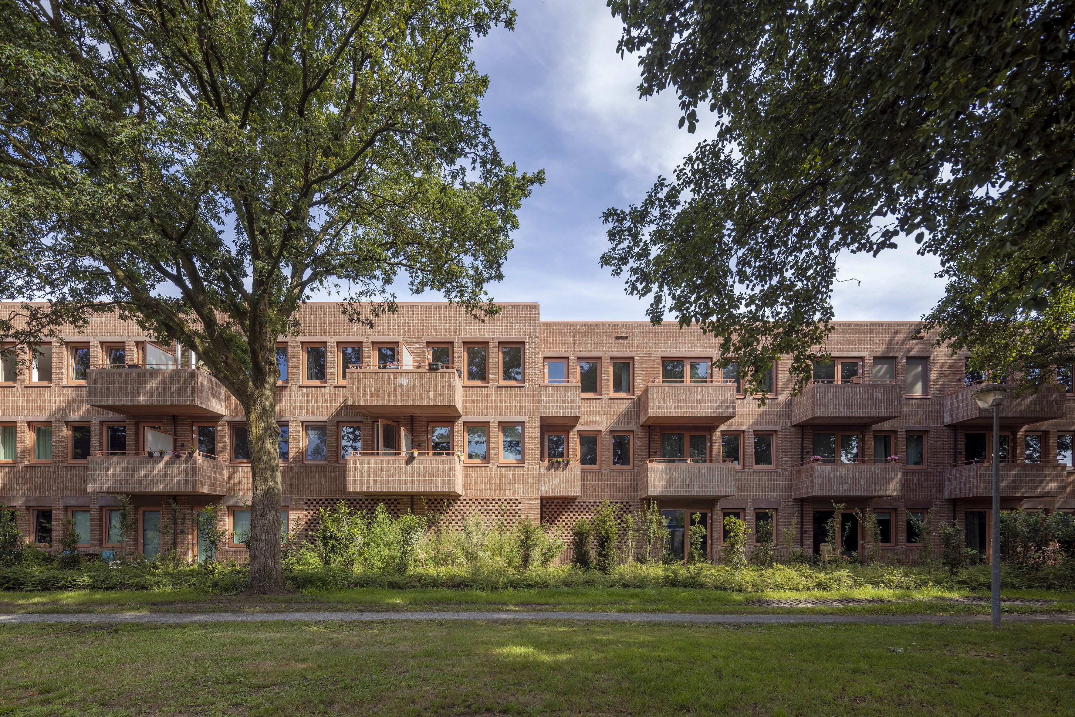 Rood bakstenen appartementengebouw met balkons, deels verborgen achter bomen.