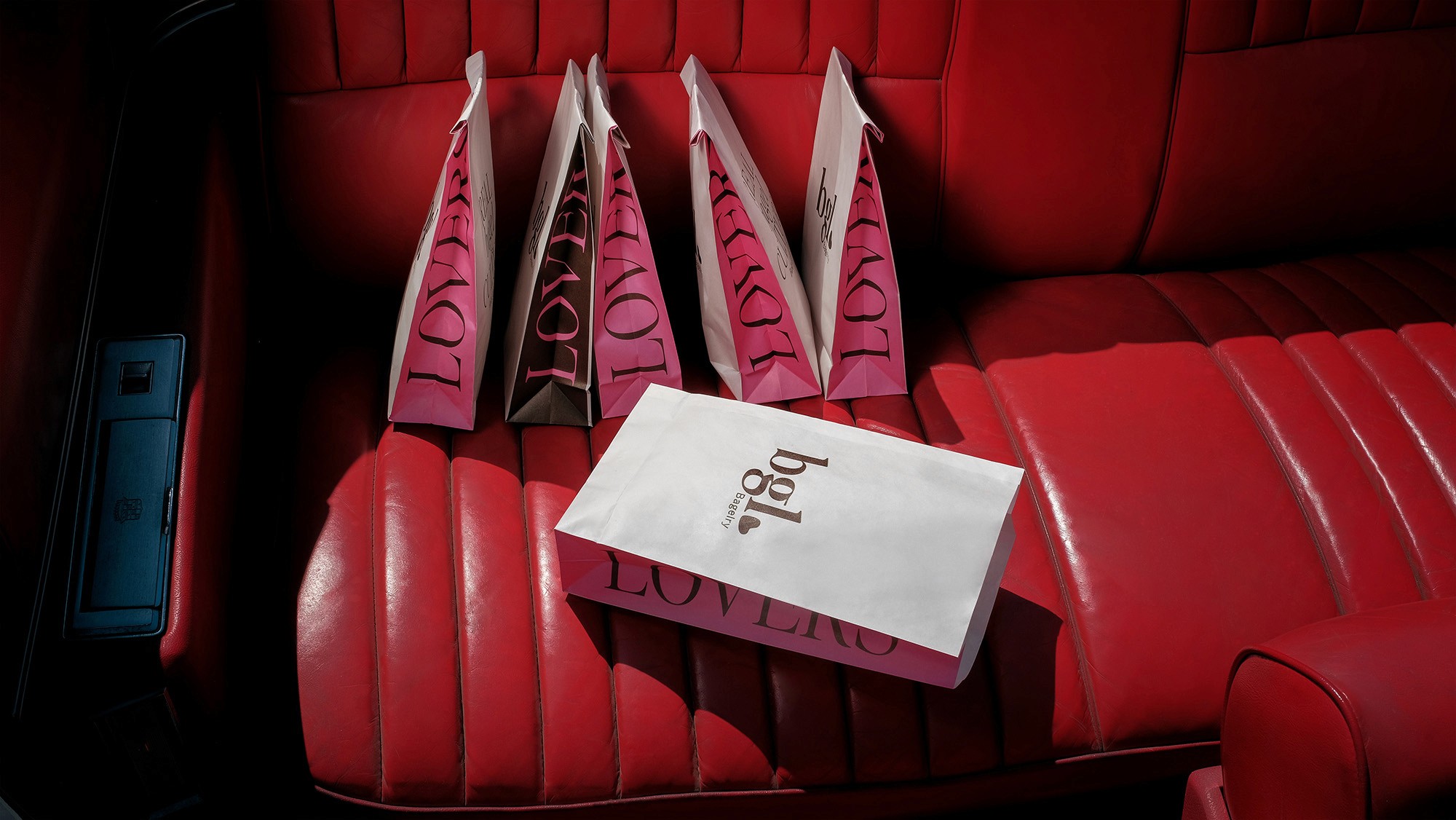 Close-up of pink "LOVE" and "bgl. LOVERS" bags sitting on a red car seat. Dramatic lighting.
