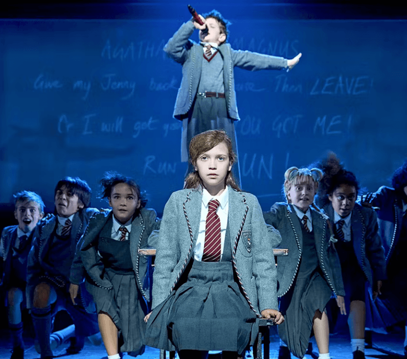 The students in their uniforms are arranged in a v on stage. Matilda is at the point. A male student stands on a desk behind her, microphone in hand. Book Matilda tickets.