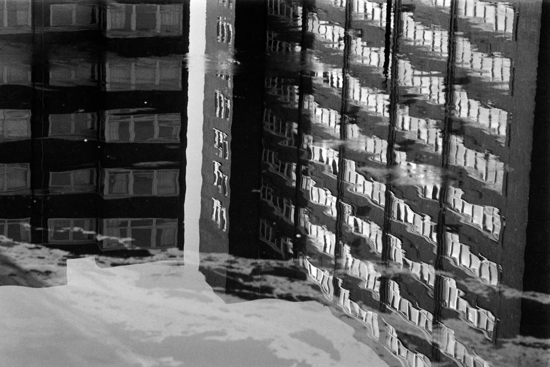 Brutalist tower block reflected in standing water, distorted windows creating fragmented architectural pattern across rippled surface, white vertical element at left edge
