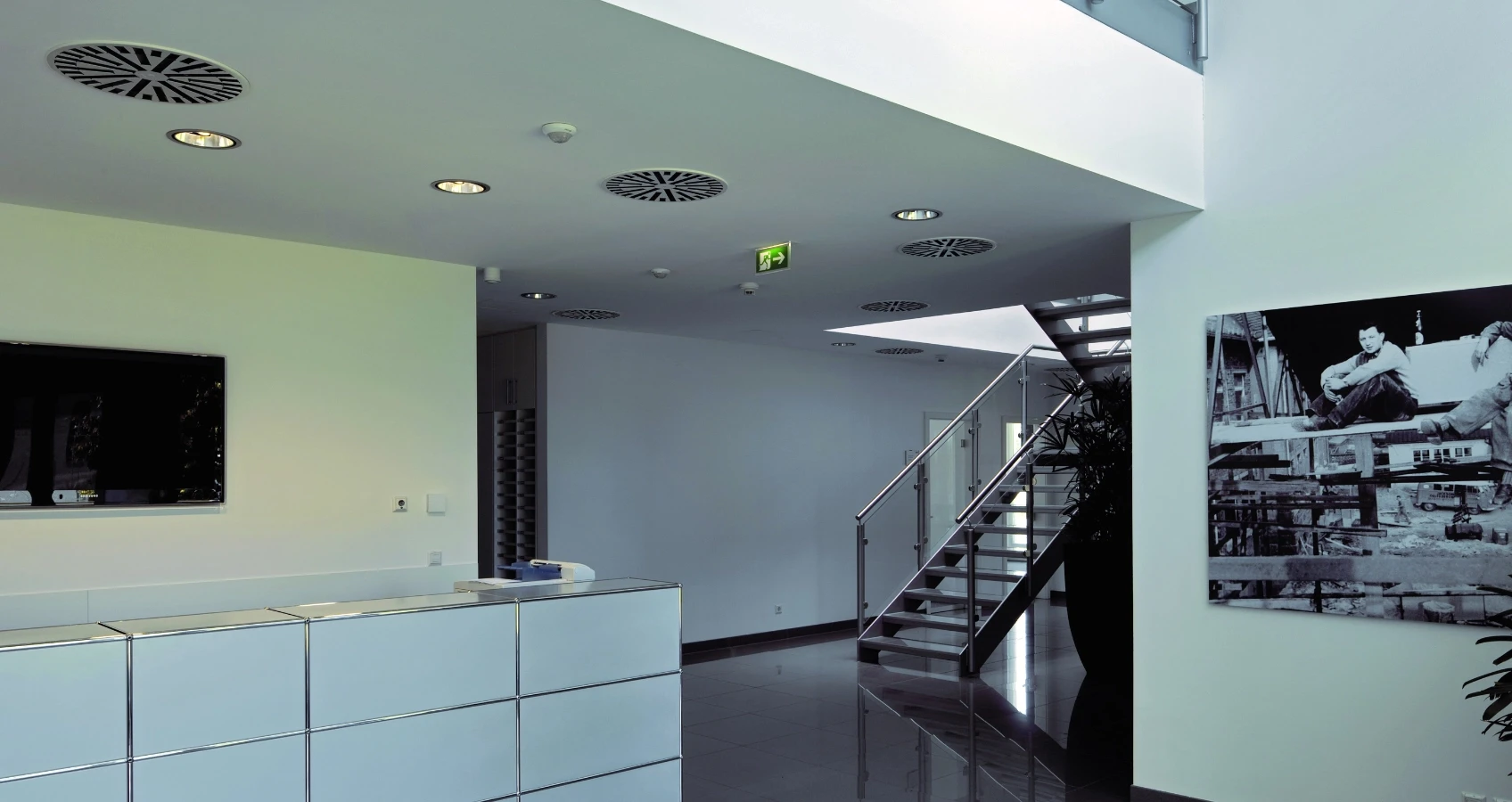 Moderner Büroempfangsbereich mit Rezeption, schwarz-weißer Wanddekoration, Treppenaufgang und Lüftungsgittern an der Decke.