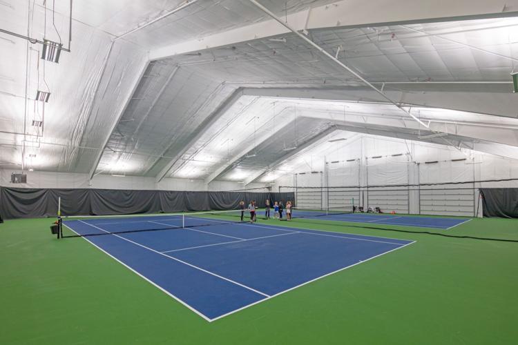 Indoor tennis court featuring a blue playing surface and green surrounding area under bright overhead lighting.