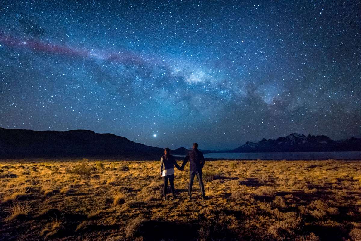 Two people stand in a grassy field at night under a vast starry sky featuring a prominent Milky Way galaxy.
