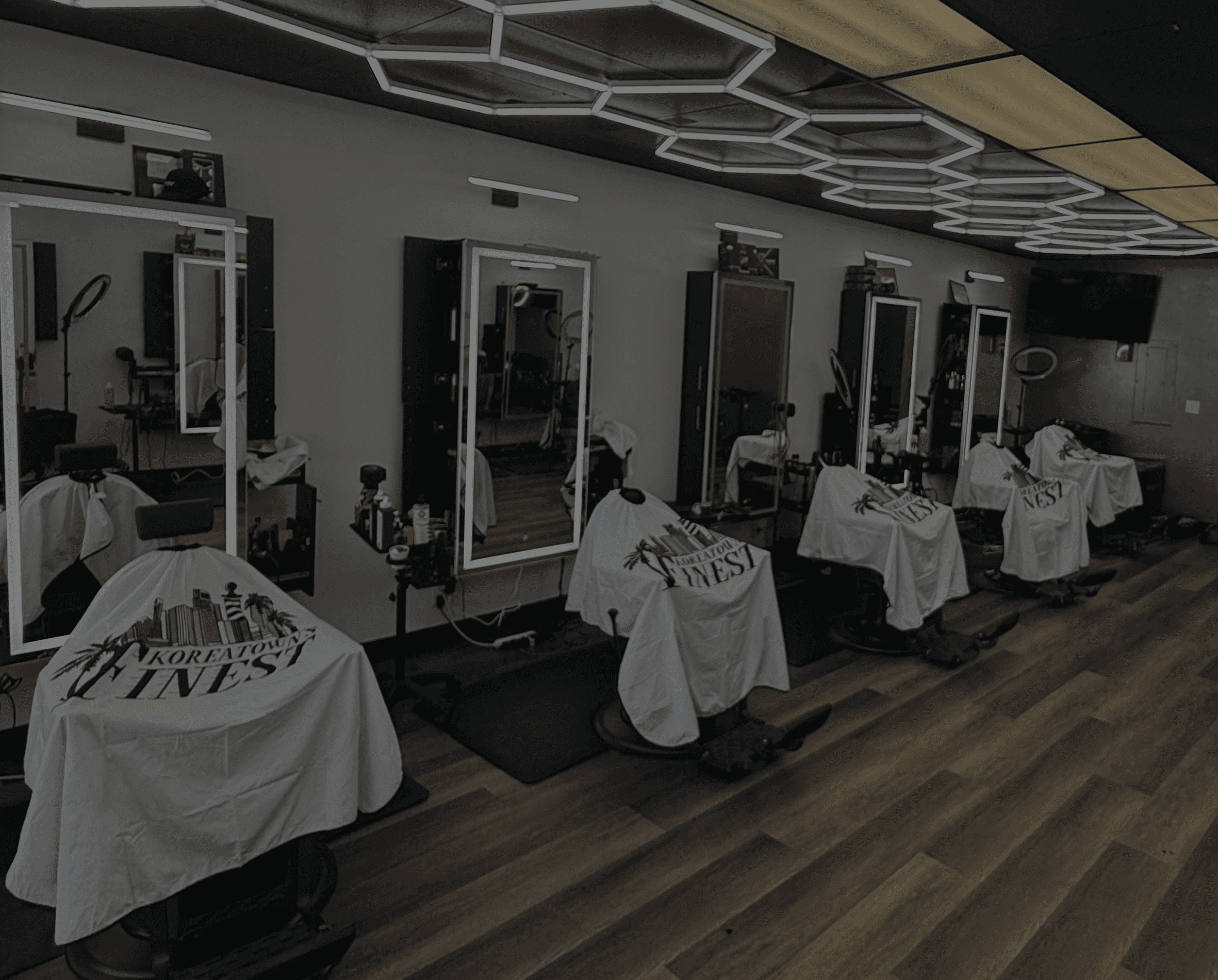 Modern barbershop interior with five barber chairs lined against a wall of mirrors, bright geometric ceiling lights, and wood-style flooring..