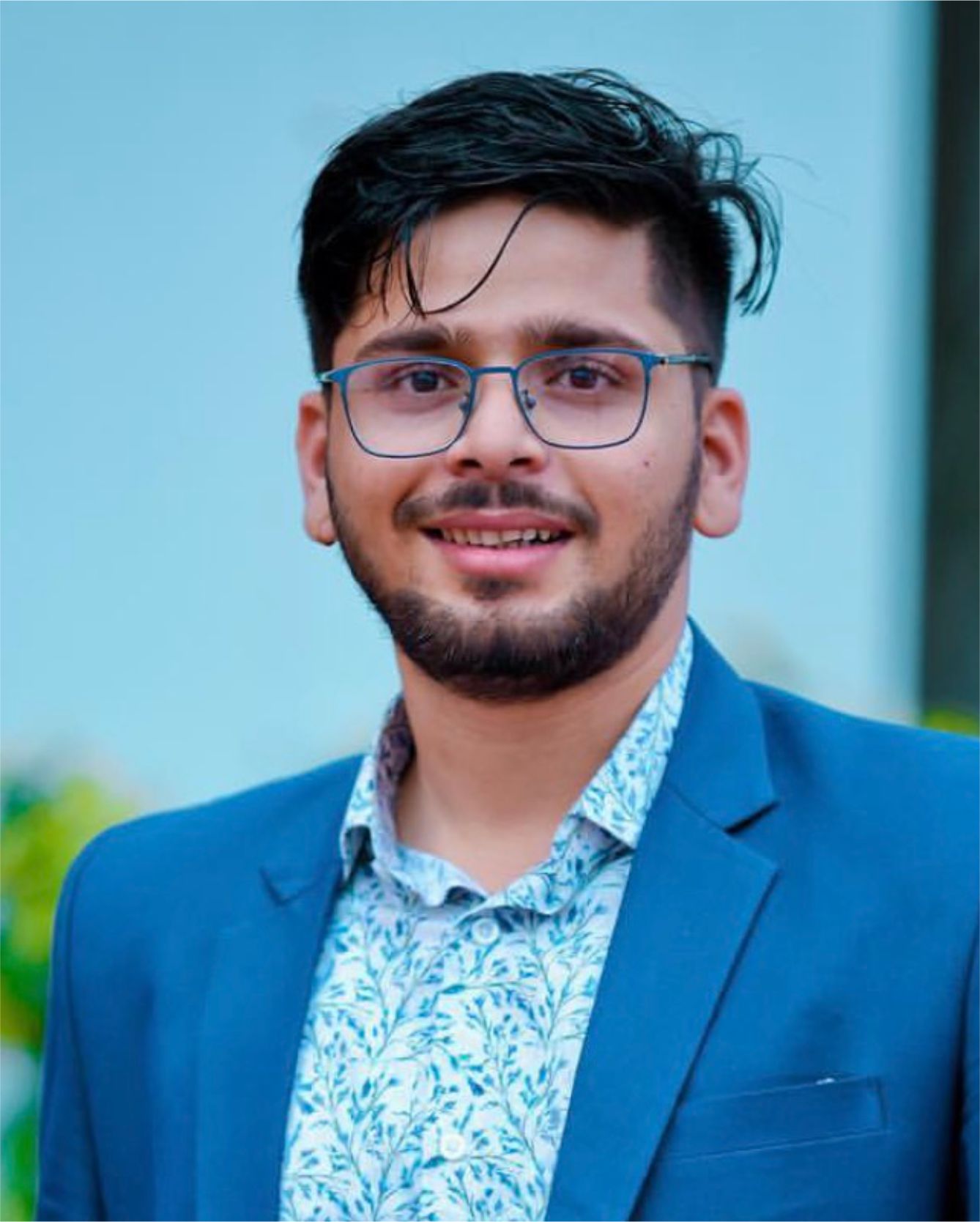 A young man wearing glasses and a blue sweater sits at a white table, flanked by a potted plant and a glowing lamp, set against a blue background.