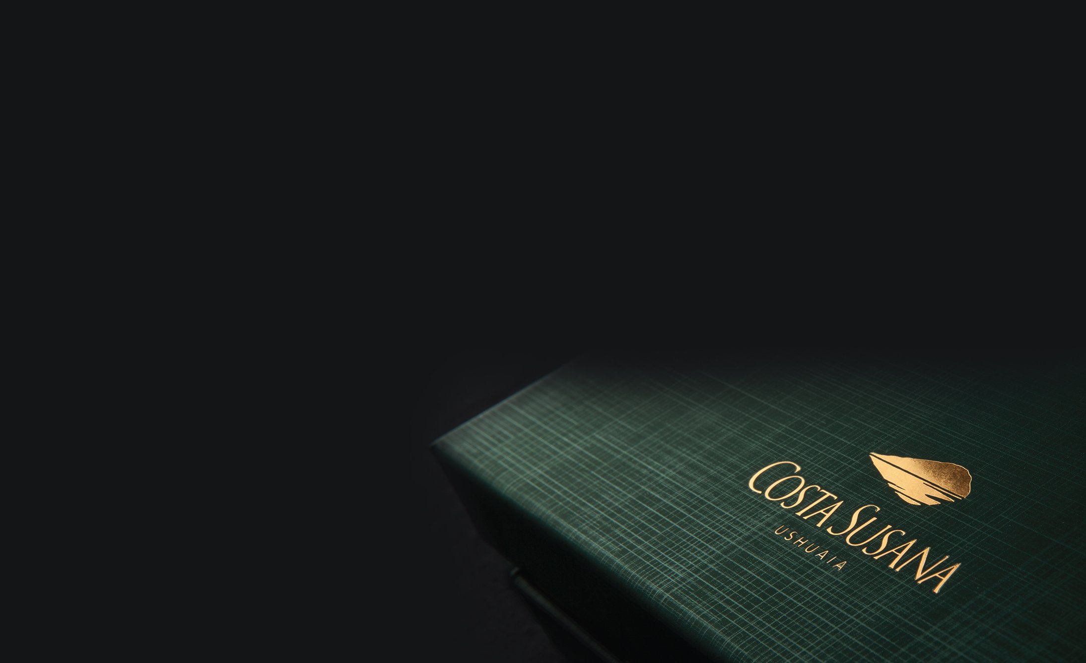 A dark green box with a textured surface is partially visible against a black background. It features gold text and a leaf logo, reading "Costa Susana Ushuaia."