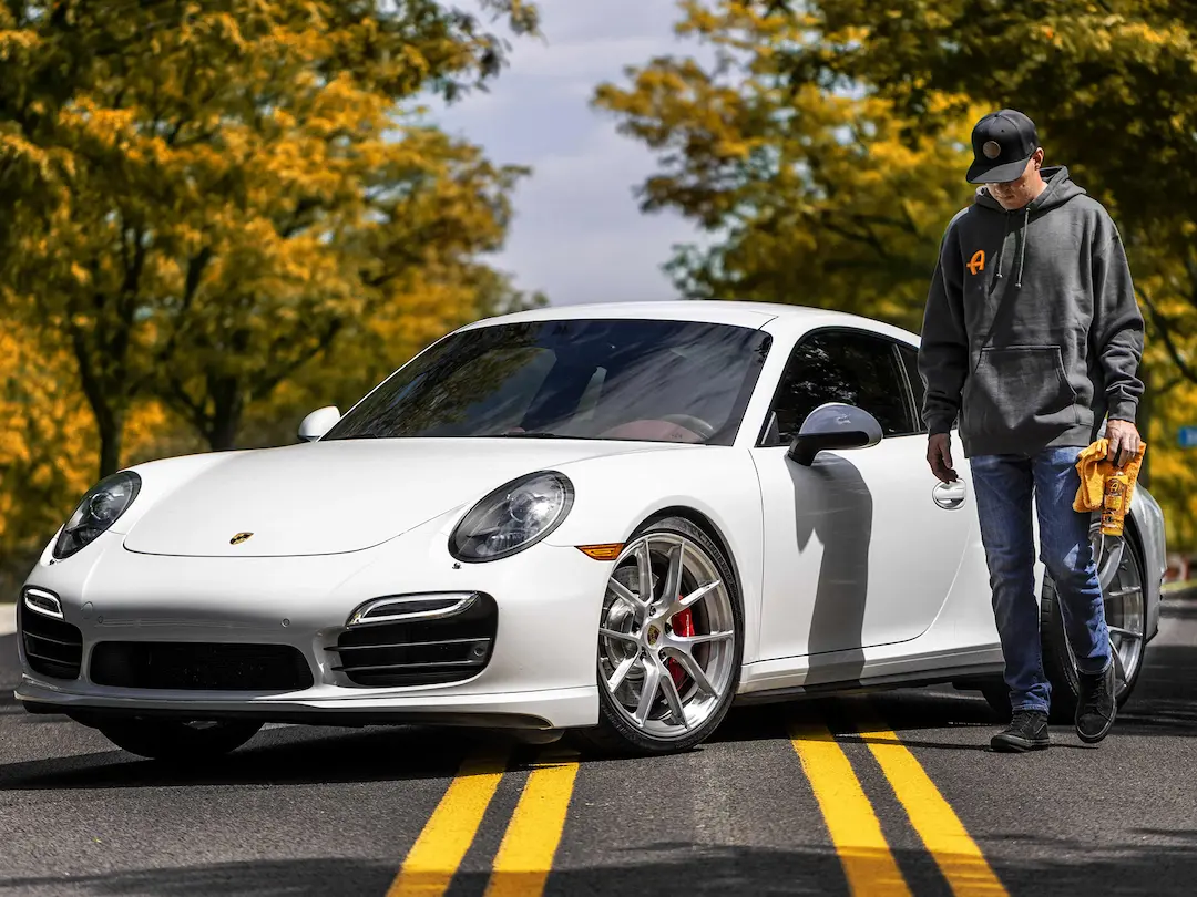 Person wearing Adams Polishes hoodie walking beside a white Porsche sports car on a tree-lined road, holding car detailing products during an autumn shoot.