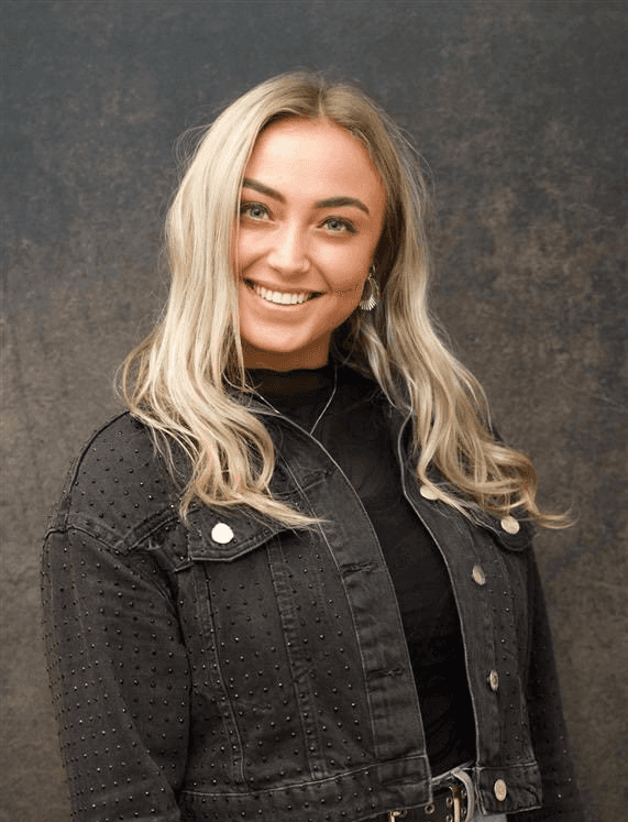 Smiling female therapist with short gray hair, wearing a brown top and a purple shawl, surrounded by a cozy background.