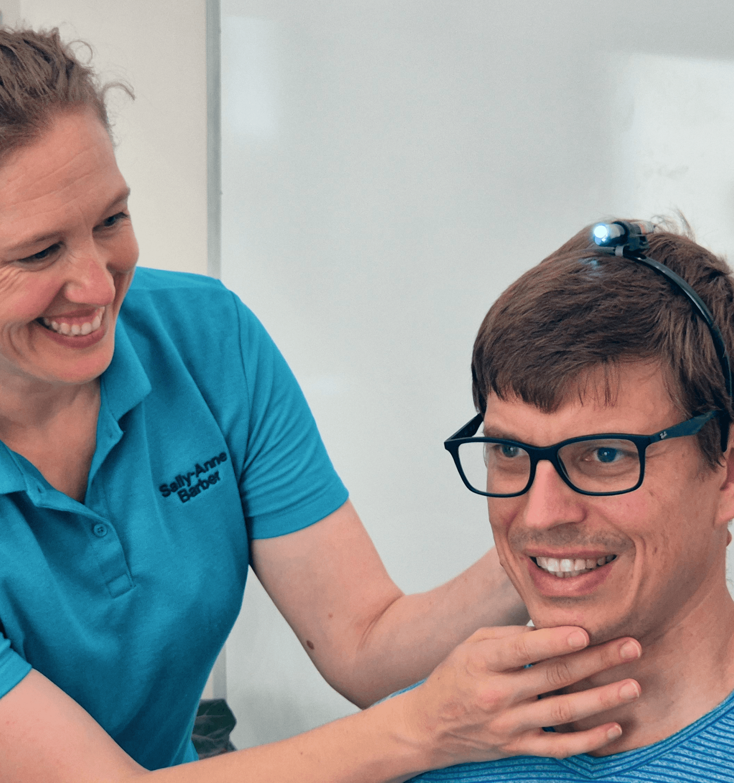 A smiling woman adjusts the chin of a young man wearing glasses, both dressed in blue shirts.