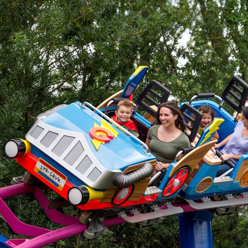 People smiling and enjoying a ride on a colorful rollercoaster with a forest backdrop.