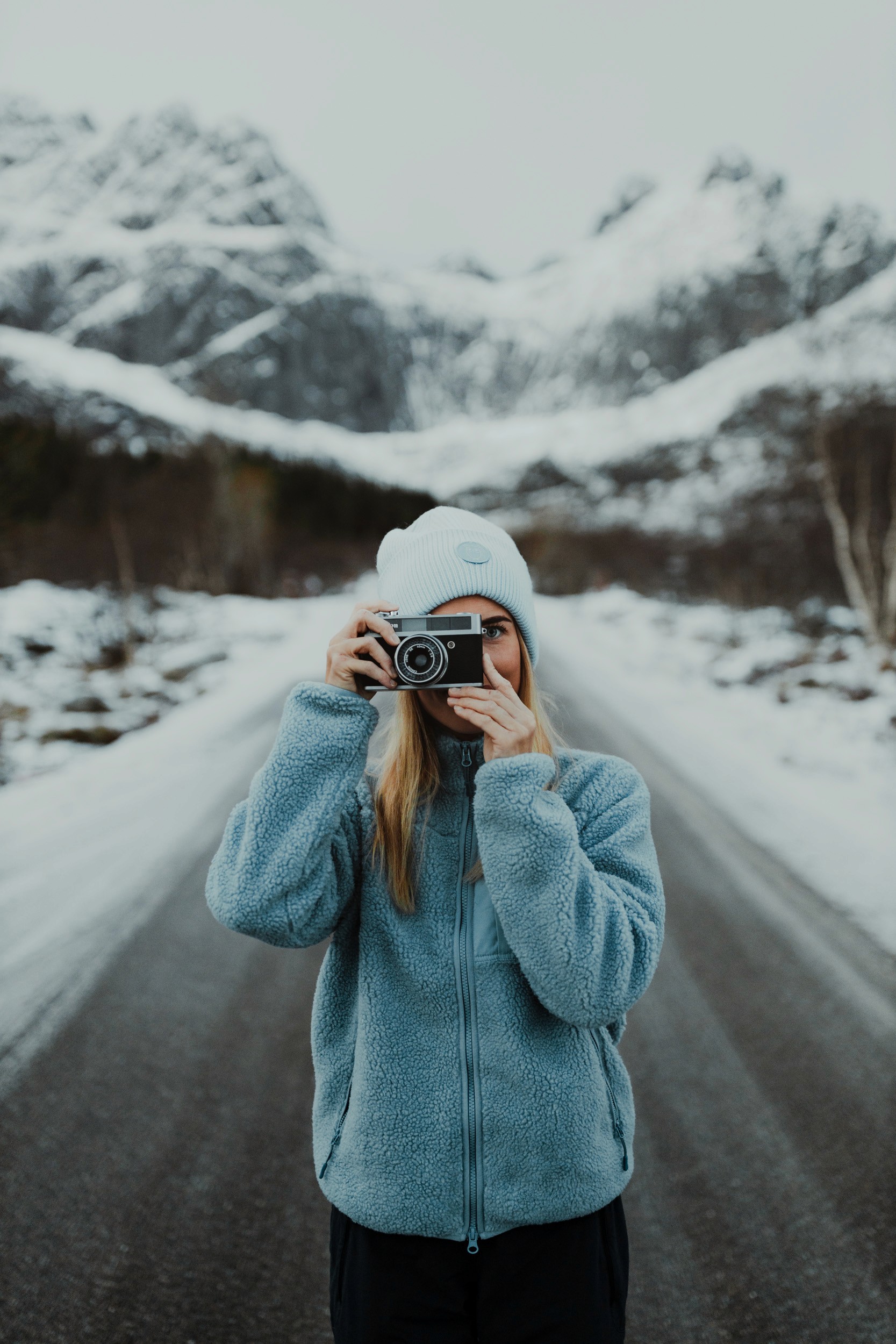 Une femme portant un bonnet et une polaire bleu prend le photographe en photo, avec des sommets enneigés derrière