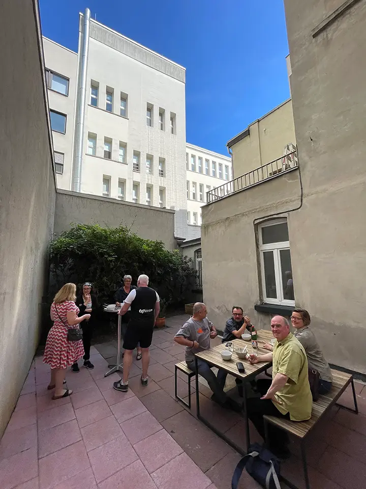 Menschen unterhalten sich in einem schmalen städtischen Innenhof mit Gebäuden im Hintergrund. Einige stehen an einem hohen Tisch, während andere an einem Holztisch sitzen und Getränke genießen. Die Szene wirkt ungezwungen und entspannt unter einem klaren blauen Himmel.