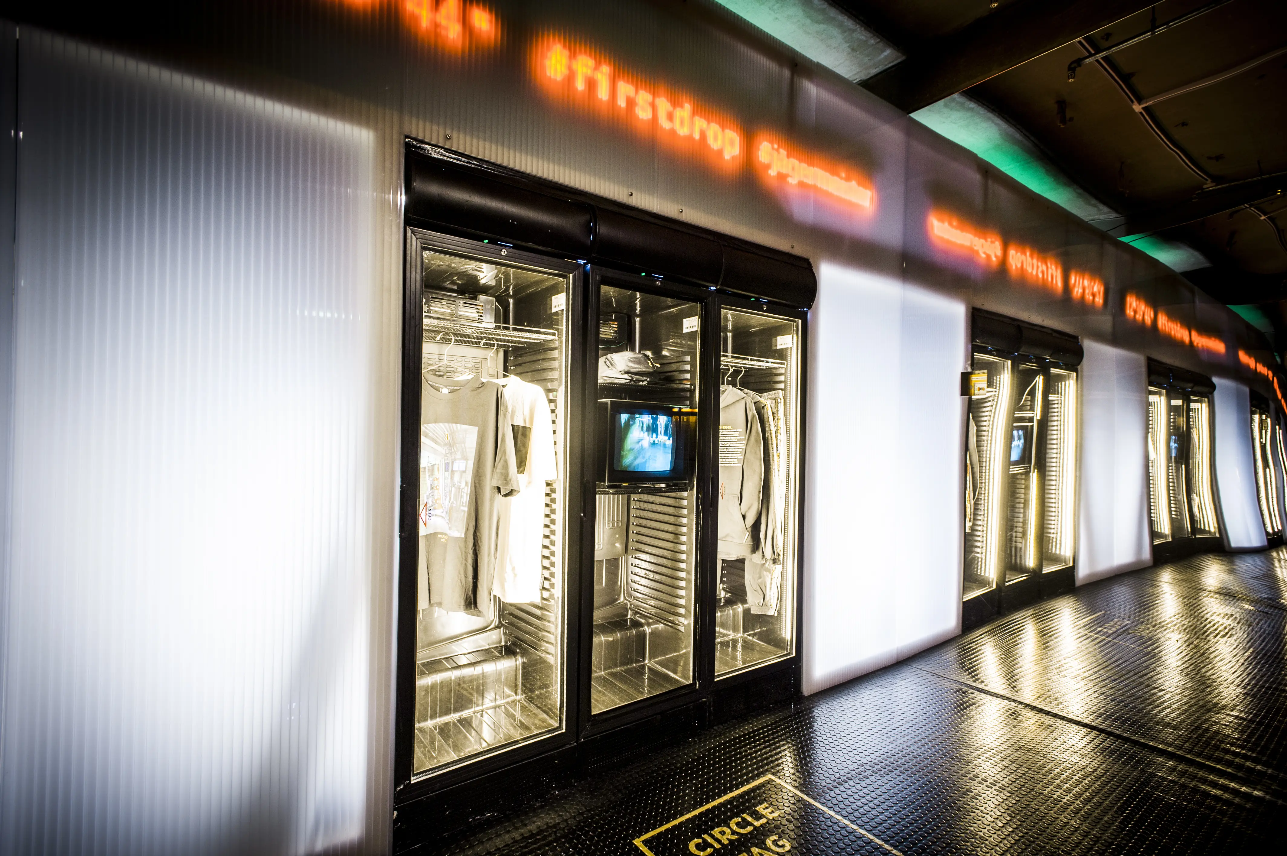 Row of backlit fridge displays with hanging streetwear and small TV screens at the JÄGERMEISTER launch, under an orange LED ticker with #firstdrop.