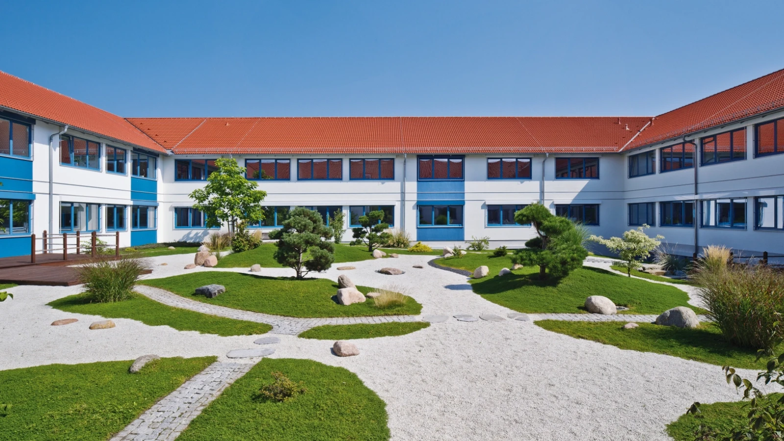 Moderne, L-förmige Architektur mit roten Ziegeldächern und blauen Fensterrahmen, die einen angelegten japanischen Zen-Garten mit Kieswegen, sorgfältig geschnittenen Bäumen und Steinen umgibt.