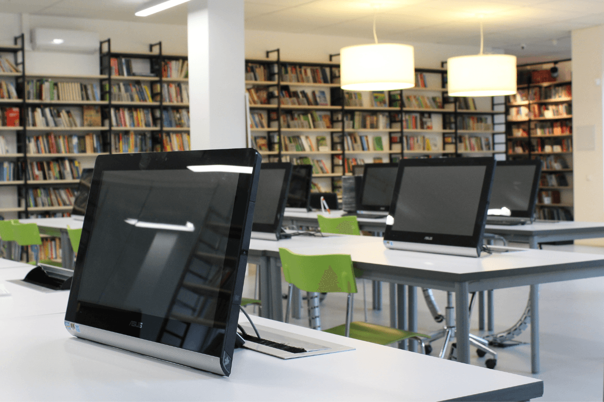 Classroom with tables with computers set up at each seat with bookcases full of books in the background and two pendant lights emitting warm glow