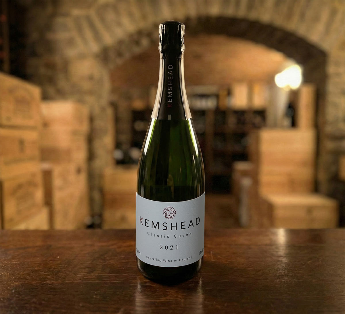 A bottle of Kemshead Classic Cuvée 2021 sparkling wine standing on a wooden counter. The bottle is dark green glass with a white label and black foil neck. The background features a rustic stone archway and stacks of wooden wine crates in a cellar setting.