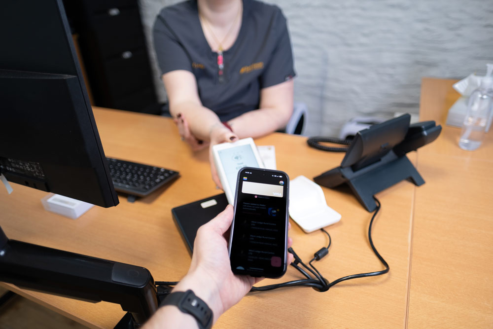A person holds a smartphone displaying a digital payment app towards a cashier's hand at a modern checkout counter, surrounded by gadgets and a wooden desk.