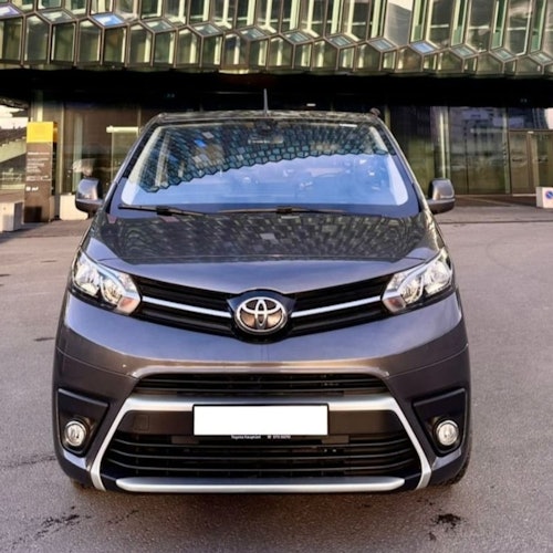 Vista frontal de una minivan Toyota gris estacionada en una calle con un moderno edificio de vidrio al fondo.