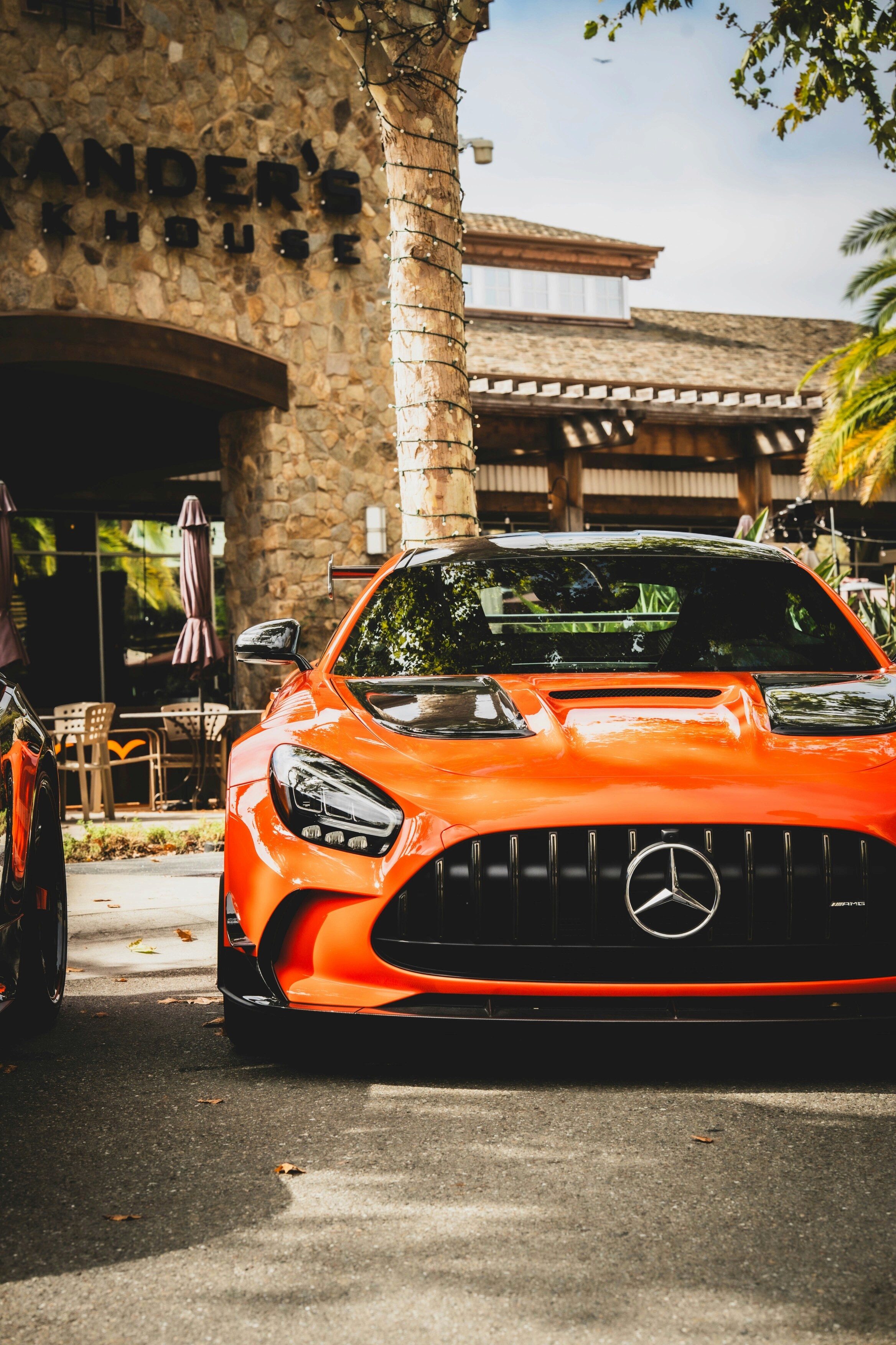 A vibrant orange sports car with a sleek, aerodynamic design is parked in front of a rustic stone building under clear blue skies, showcasing stylish curves and a luxurious emblem, surrounded by lush greenery and a casual dining setting.