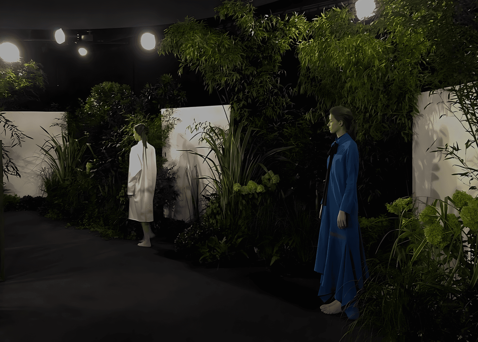 Deux mannequins en robes minimalistes, l'une en blanc et l'autre en bleu, posent dans un décor végétal luxuriant et immersif, éclairé par des projecteurs dans un espace intérieur sombre.