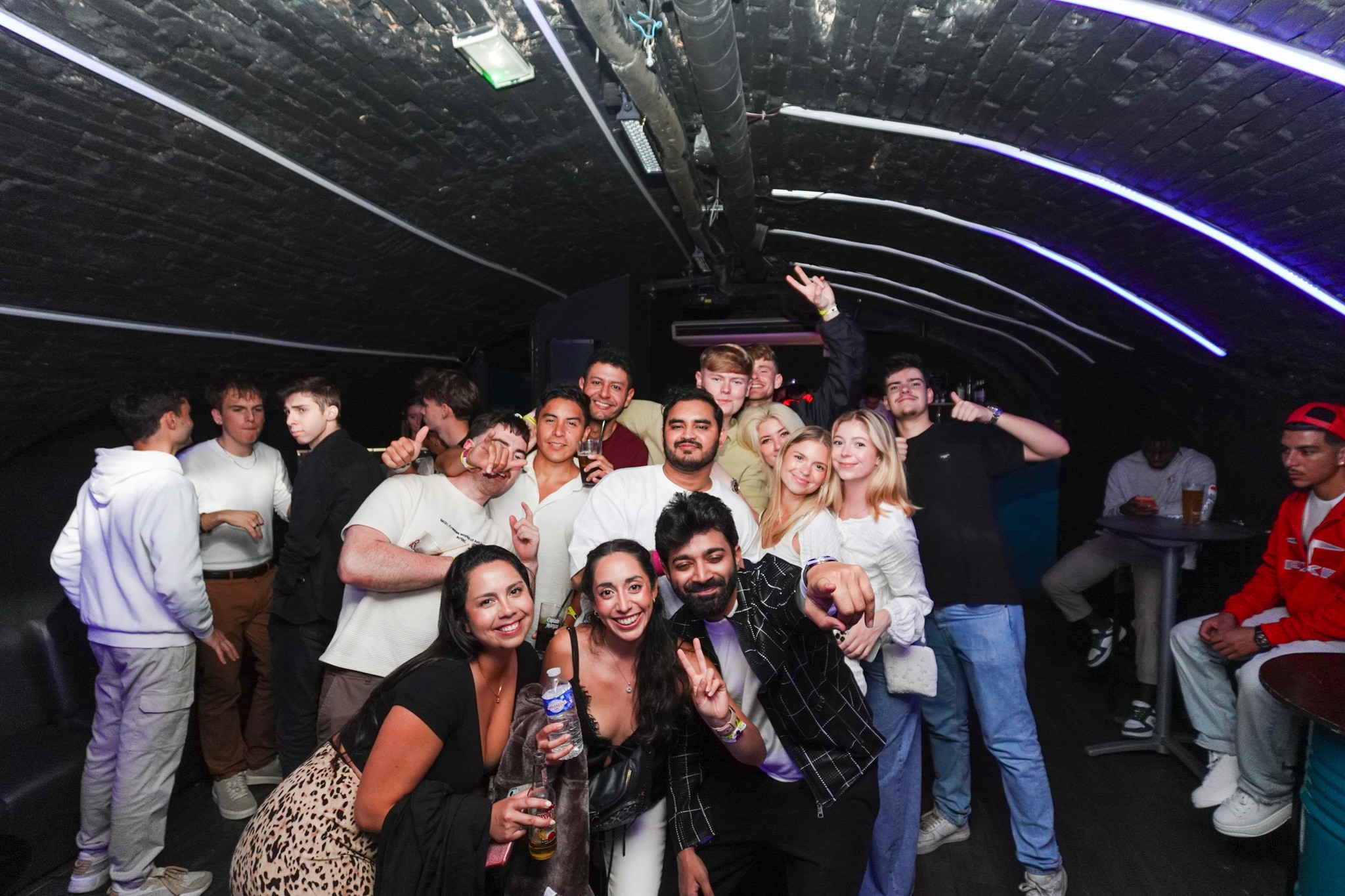 Large group of young partygoers posing together inside a packed underground bar during a bar crawl in Nice celebrating with drinks hand signs and big smiles capturing the high energy social vibe and international nightlife atmosphere of the French Riviera