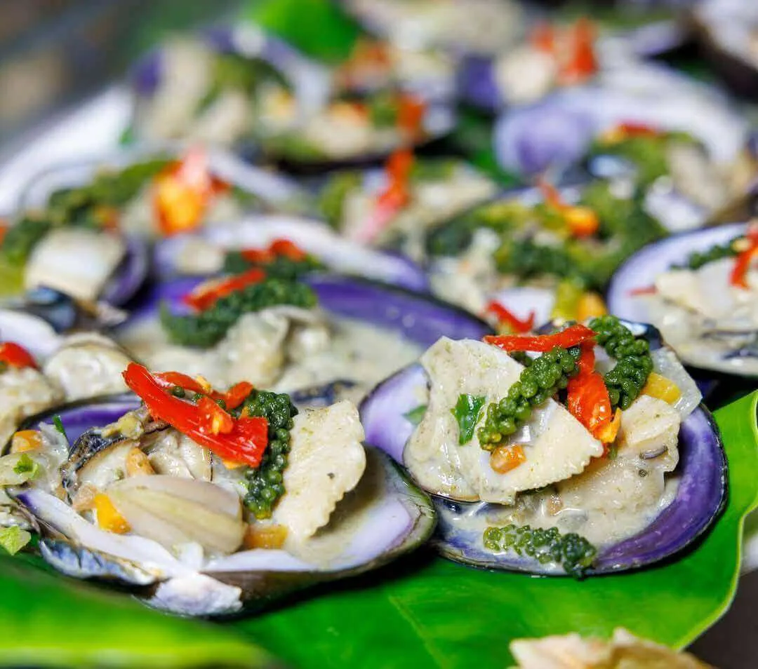 Close-up of cooked shellfish in purple shells, topped with creamy sauce, green peppercorns, and red chili slices on a leaf.