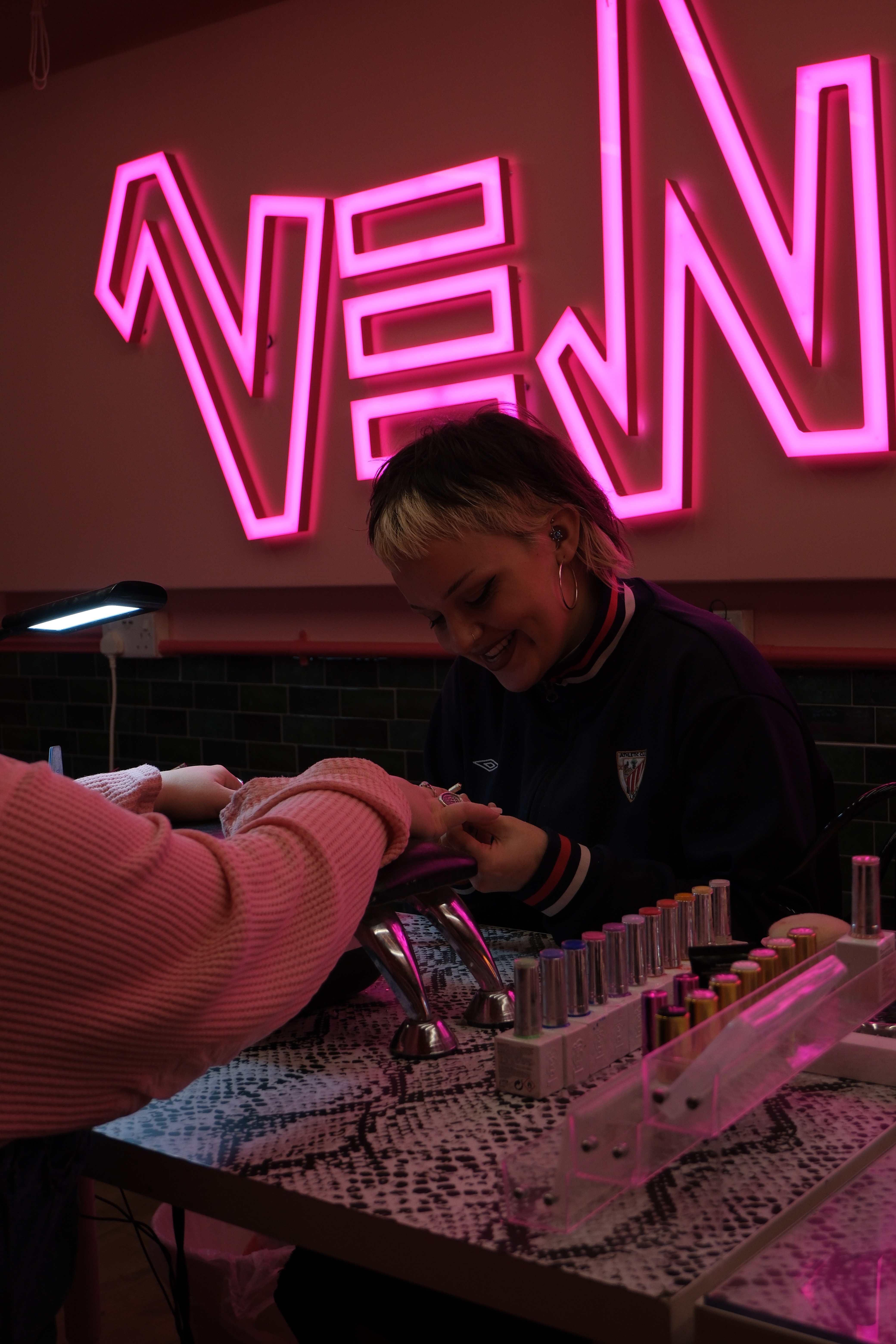 A technician looking down, concentrating on the final details of a client's set, with professional gel polish bottles lined up in the foreground.