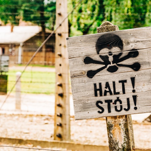 Weathered wooden sign with a skull and crossbones and the text "HALT! STÓJ!" in front of a barbed wire fence and building.