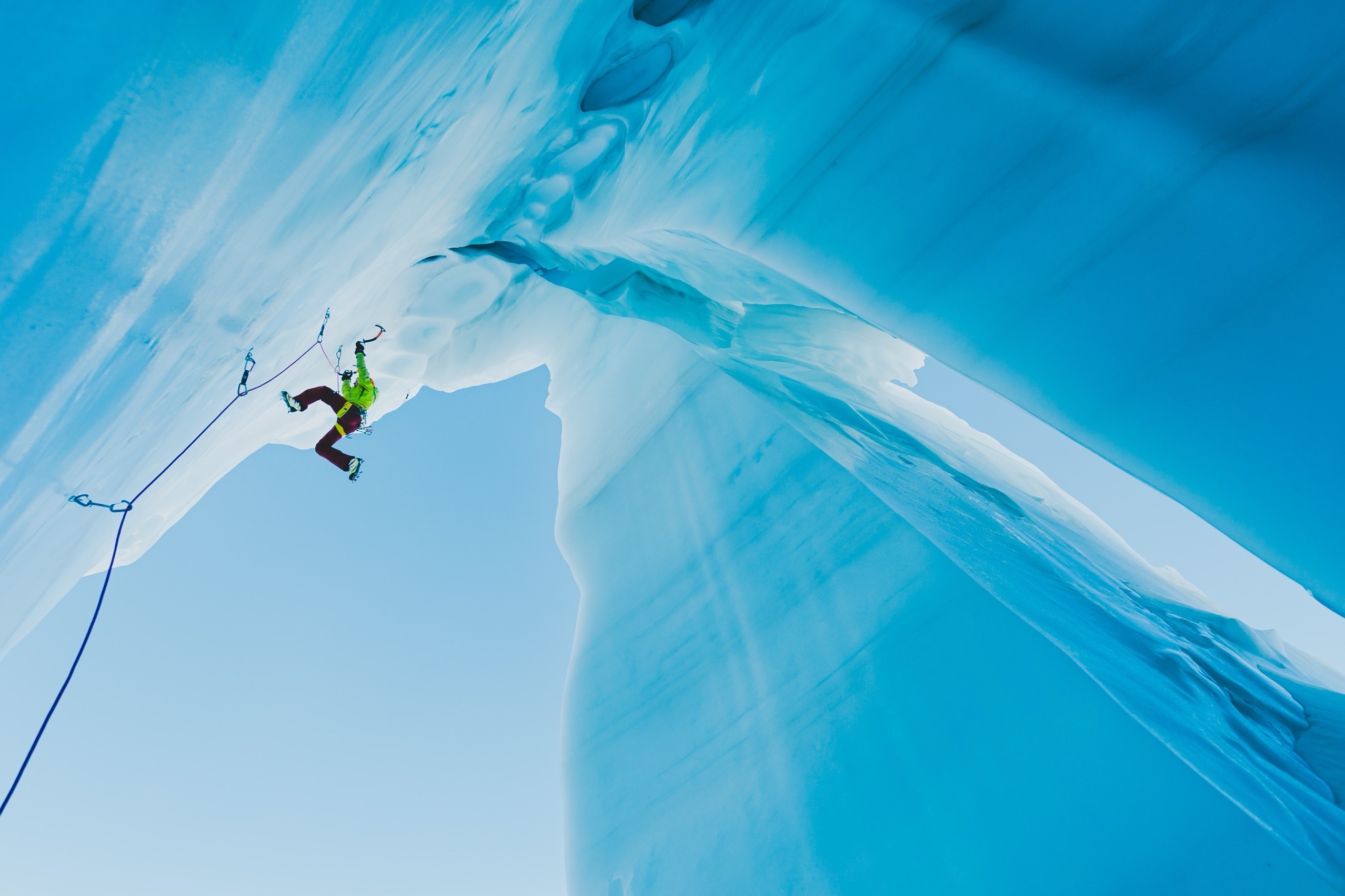 Ein Eiskletterer hängt unter dem Überhang eines blauen Gletschers, ein Eisgerät über ihm gesetzt, das Seil läuft am Eis hinunter.