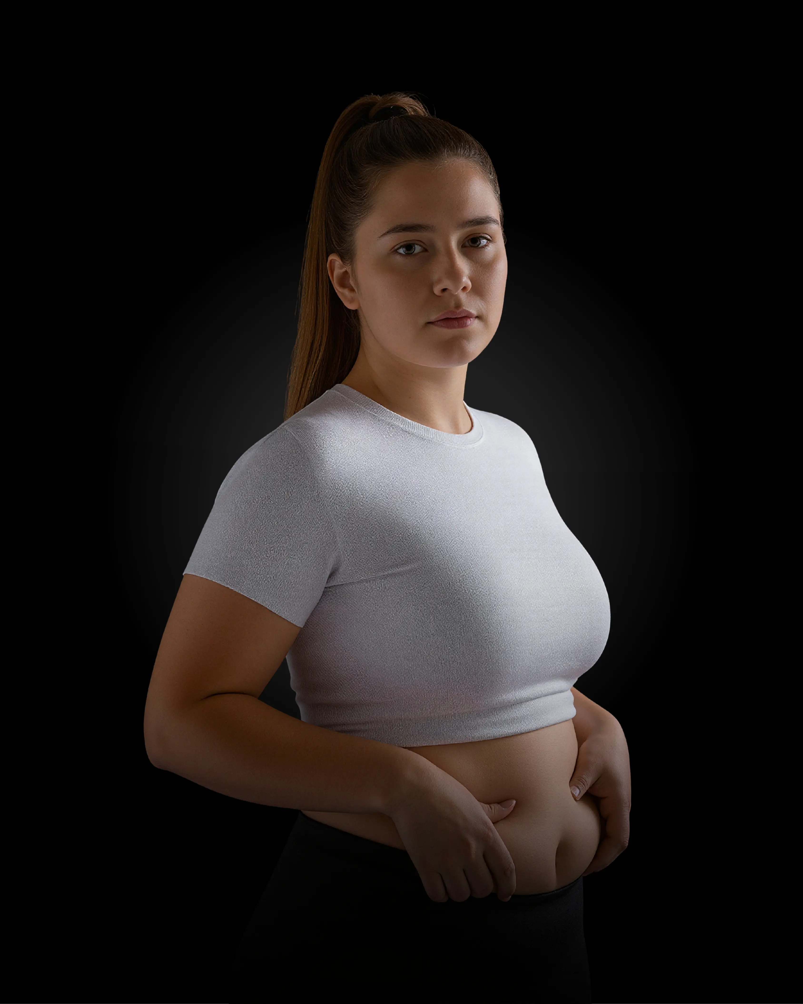 Woman in white crop top and black pants, looking at the camera, touching her belly.