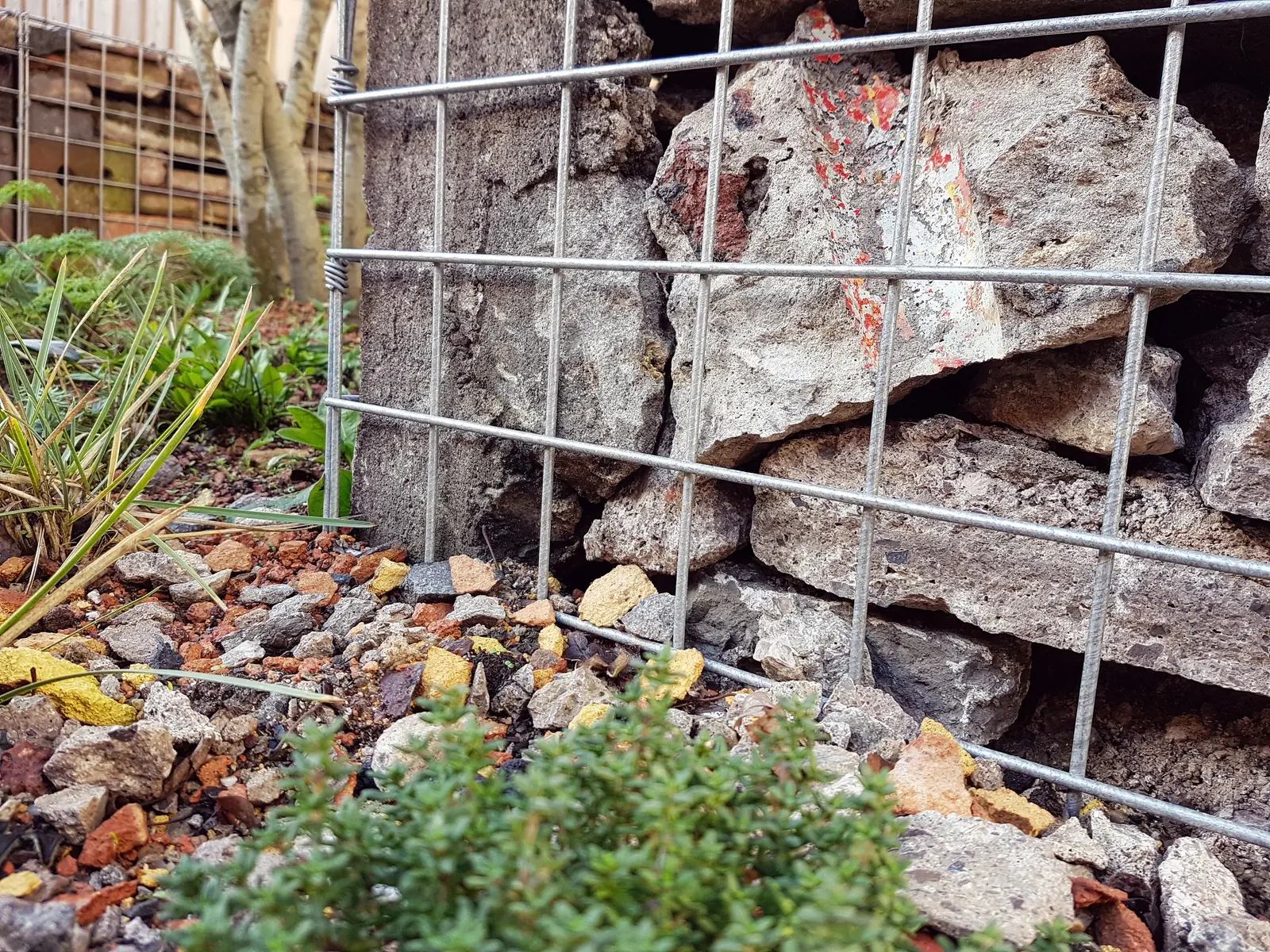 Weathered stones and bricks with greenery growing nearby, partially covered by a rusty metal grid.