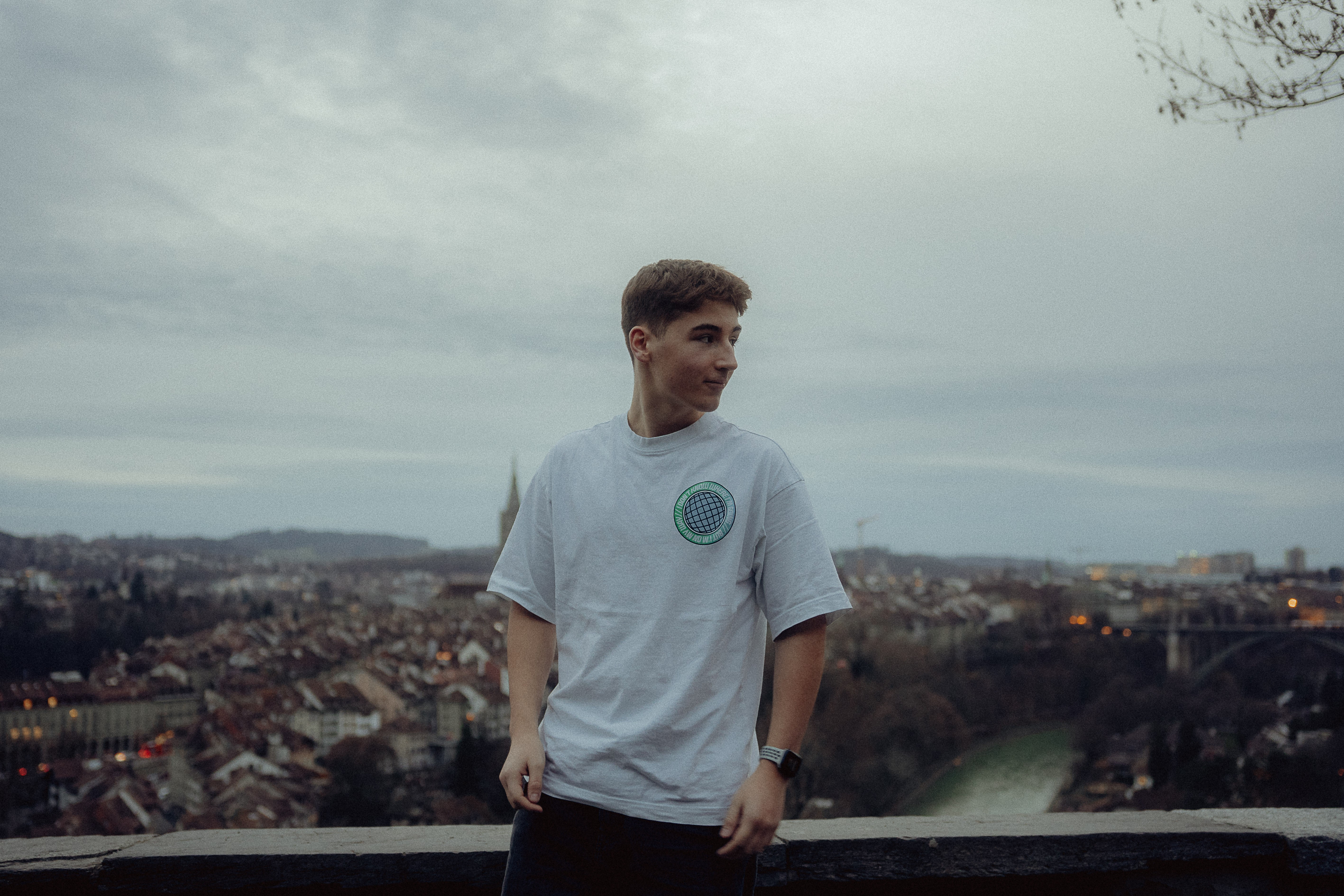 A young person stands outdoors, overlooking a cityscape under a cloudy sky, wearing a white shirt.