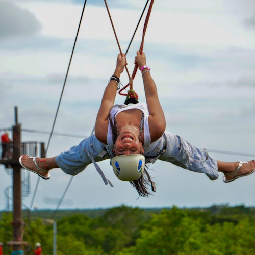 Person in a helmet and harness upside down on a zipline, smiling and spreading their arms and legs. Green trees in the background.