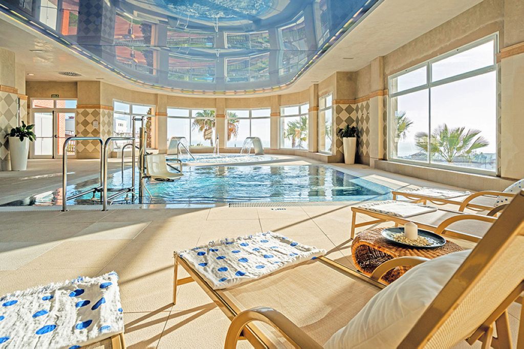 Indoor swimming pool facility with heated water, wooden decking, large curved windows with natural light, modern wellness center design, and comfortable amenities