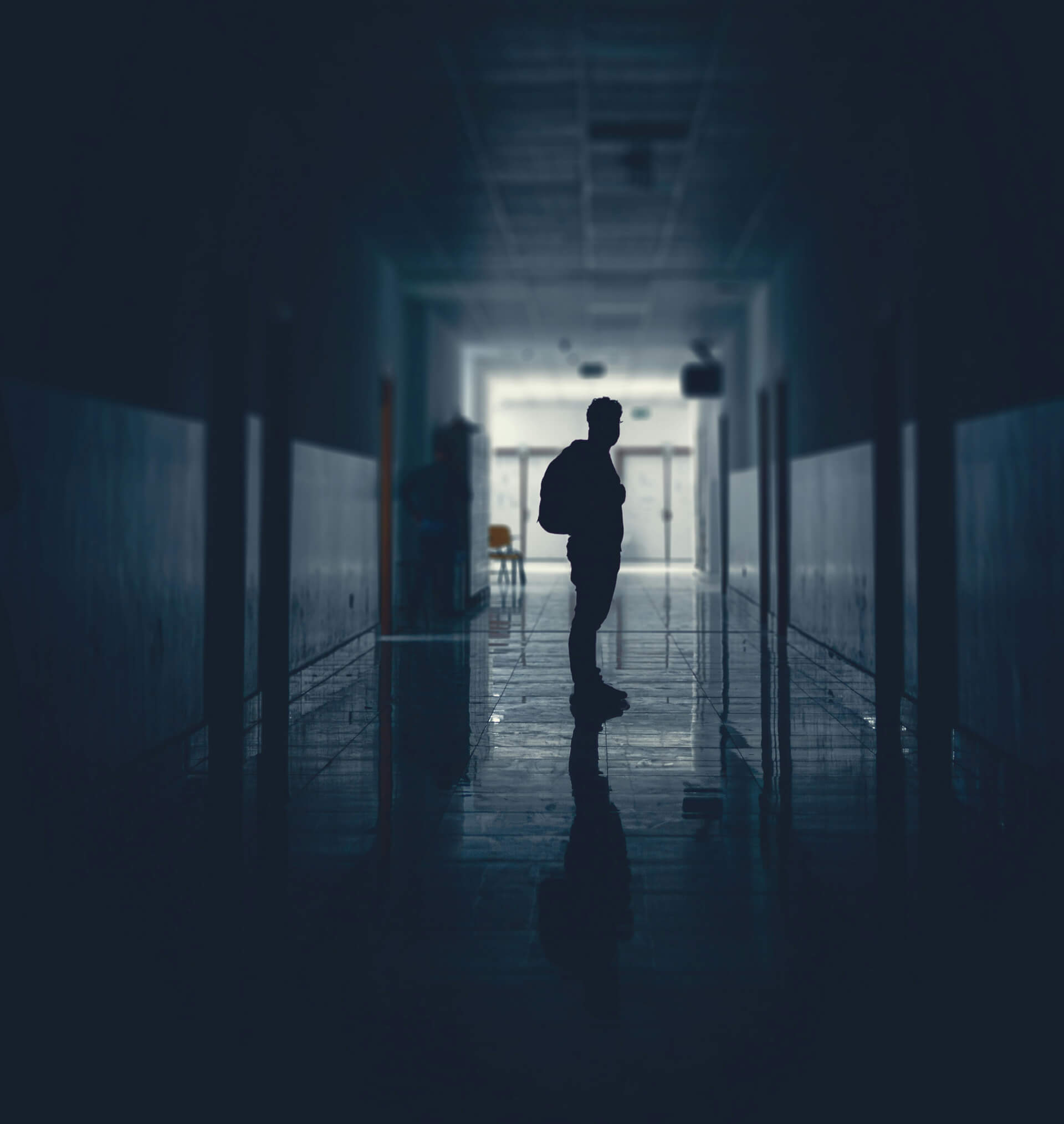 A silhouette of a person with a backpack stands alone in a dimly lit hallway, illuminated faintly by light coming from a doorway at the far end. The reflective floor mirrors the figure, adding to the moody, contemplative atmosphere.