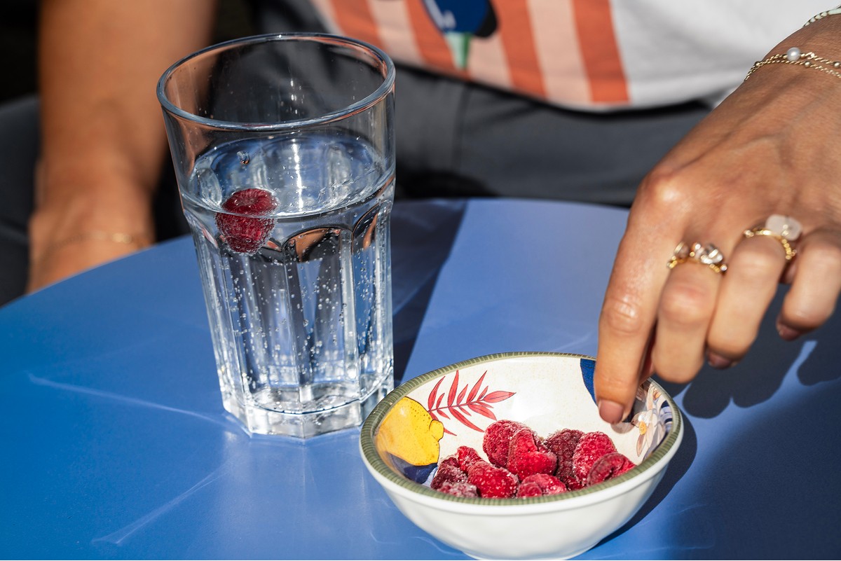A person reaches for fresh raspberries from a decorative bowl beside a glass of water with raspberries floating, placed on a vibrant blue surface, promoting a sense of wellbeing and healthy living.