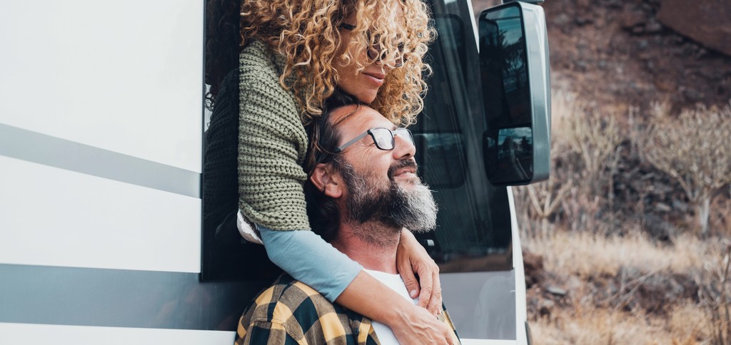 A happy couple enjoys a moment together by their RV, with the woman leaning out of the window to hug the man, symbolizing love, adventure, and the freedom of road travel.