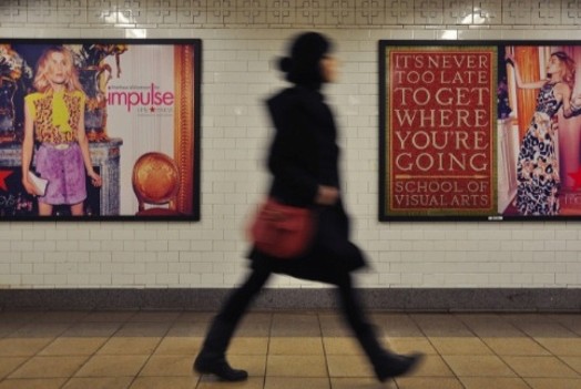 A photo of a person walking by poster of a woman and another one with a tile pattern metro direction sign that says: It's never too late to get where you're going. School of Visual Arts.