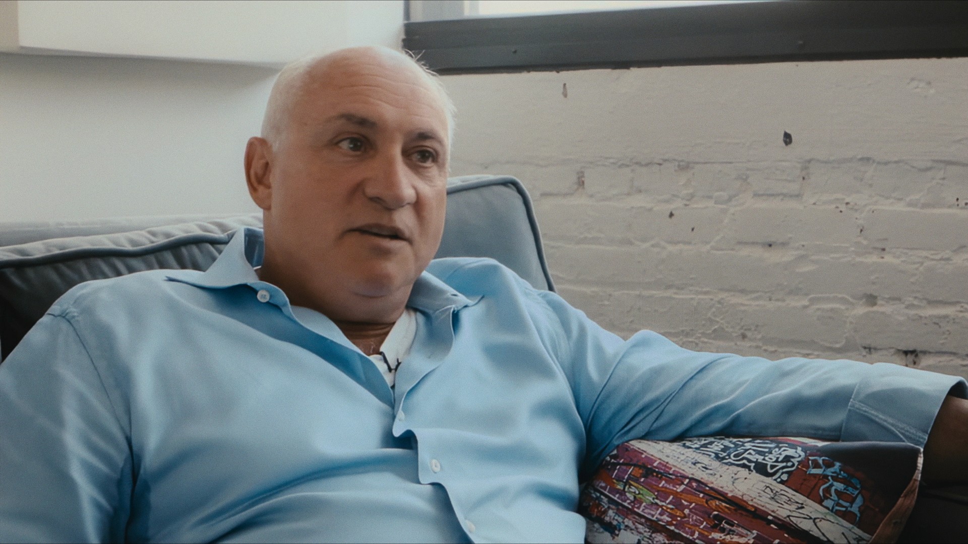 A man in a light blue shirt on a sofa, interviewed against a white brick wall, Maddman