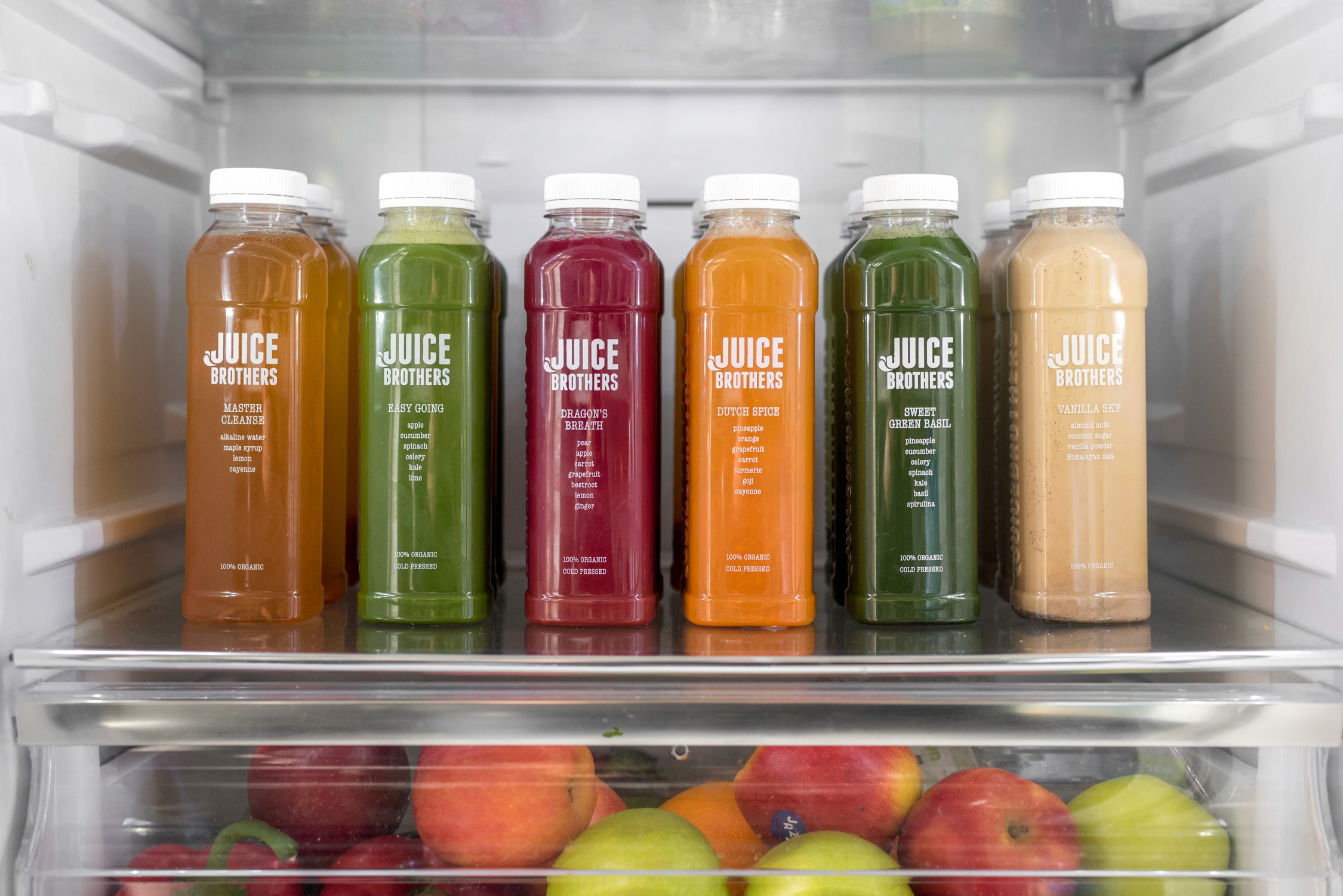 Brightly lit refrigerator shelves display colorful rows of juice bottles alongside fresh, vibrant fruits in a clear crisper drawer.