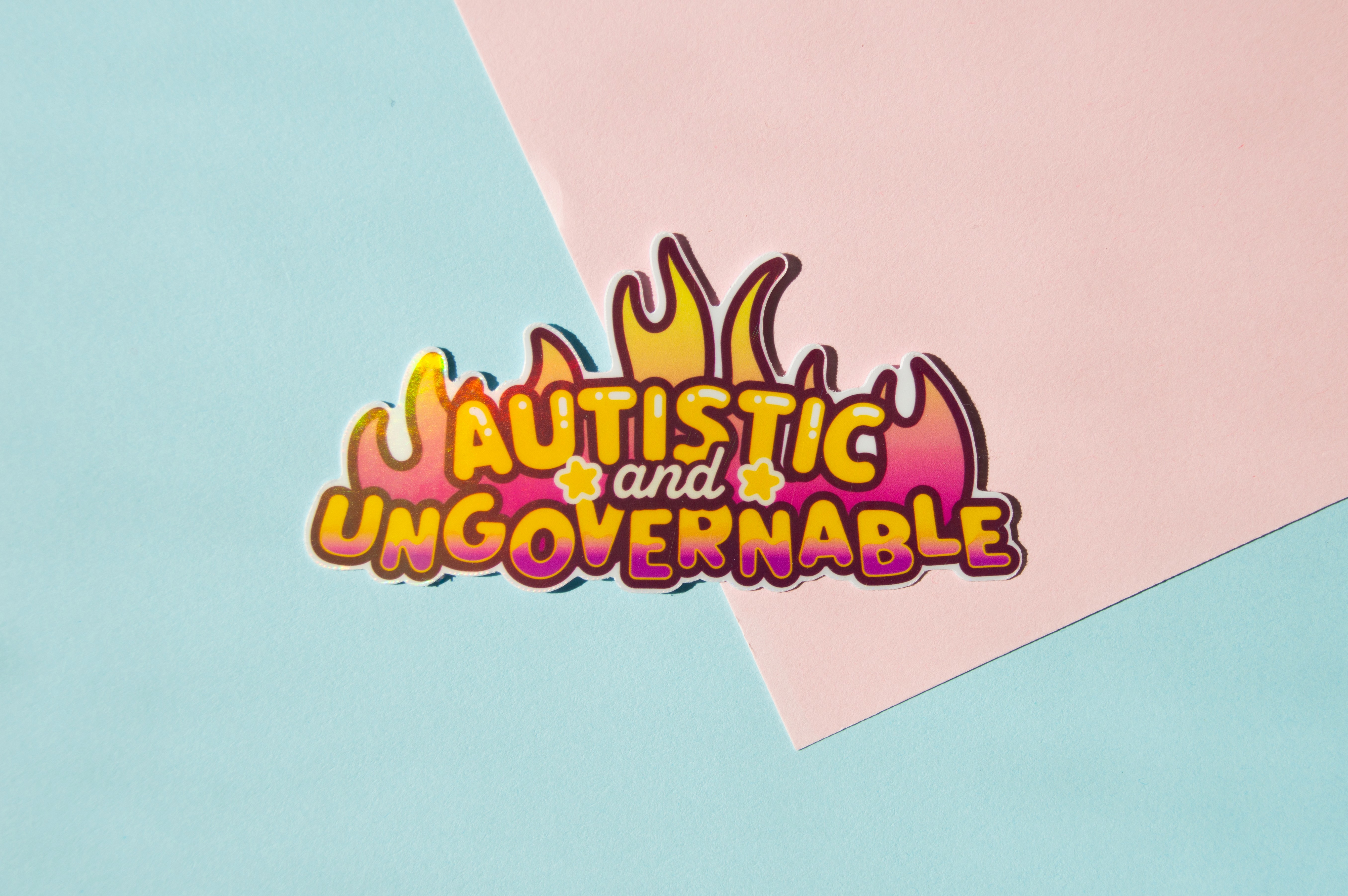 A sticker that reads "Autistic and Ungovernable" in yellow letters, turning pink at the bottom. Behind it are pink and yellow flames.