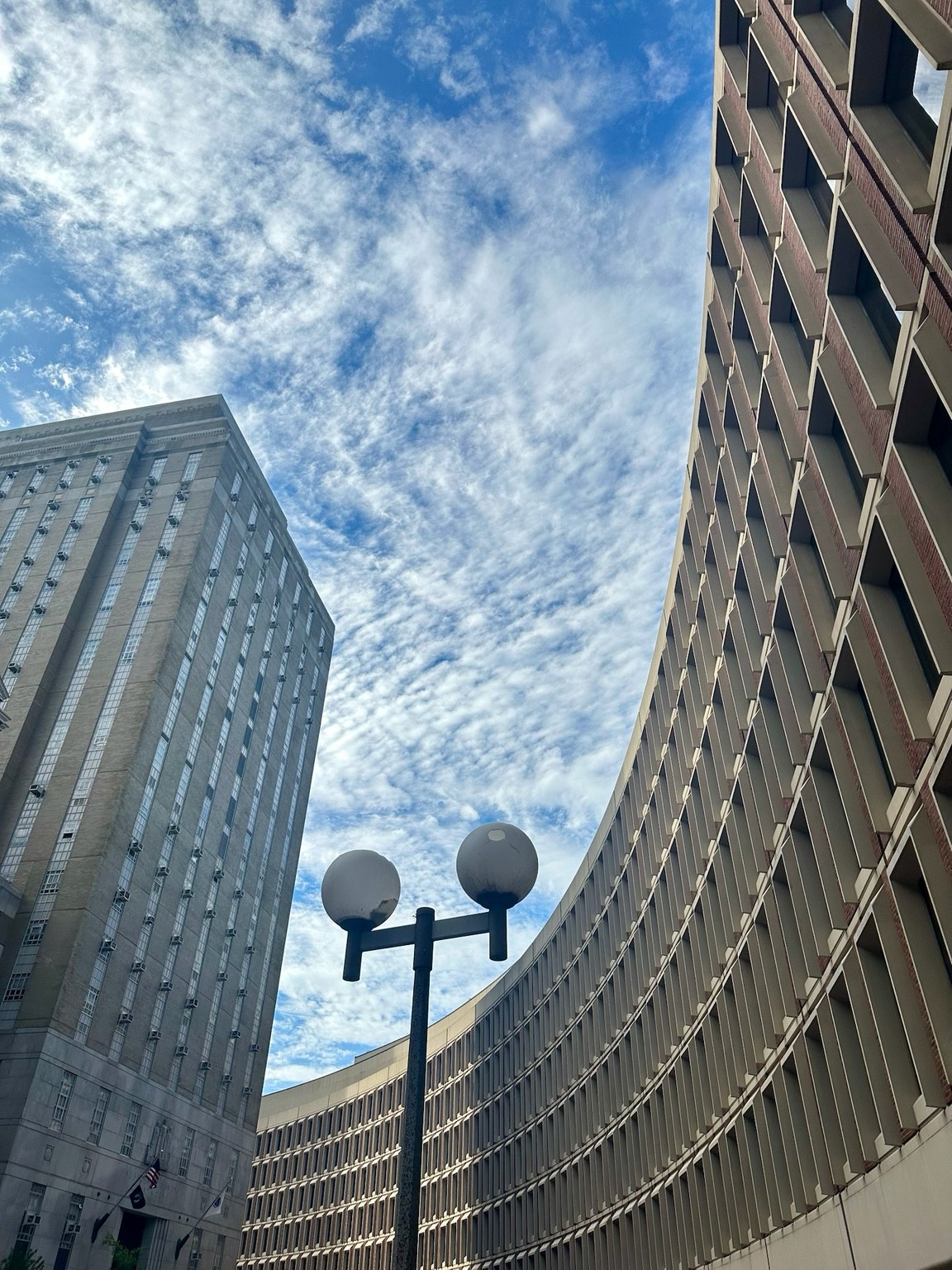 A curved grid of windows beside a tall stone building and twin globe streetlamp beneath rippled clouds.