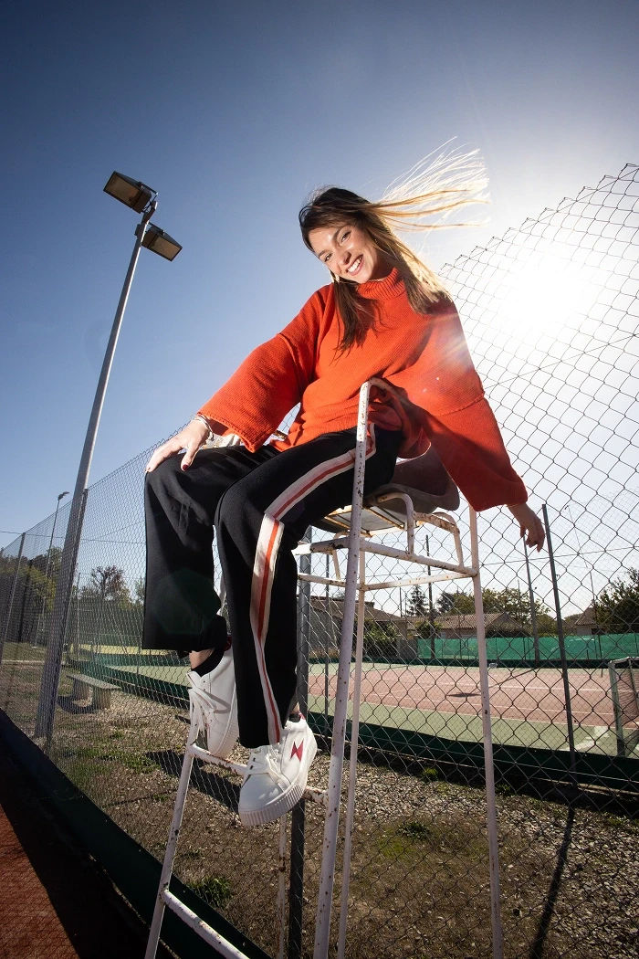 Une femme avec un pantalon noir et un pull orange. Elle est assise sur la chaise de l'arbitre d'un court de tennis.