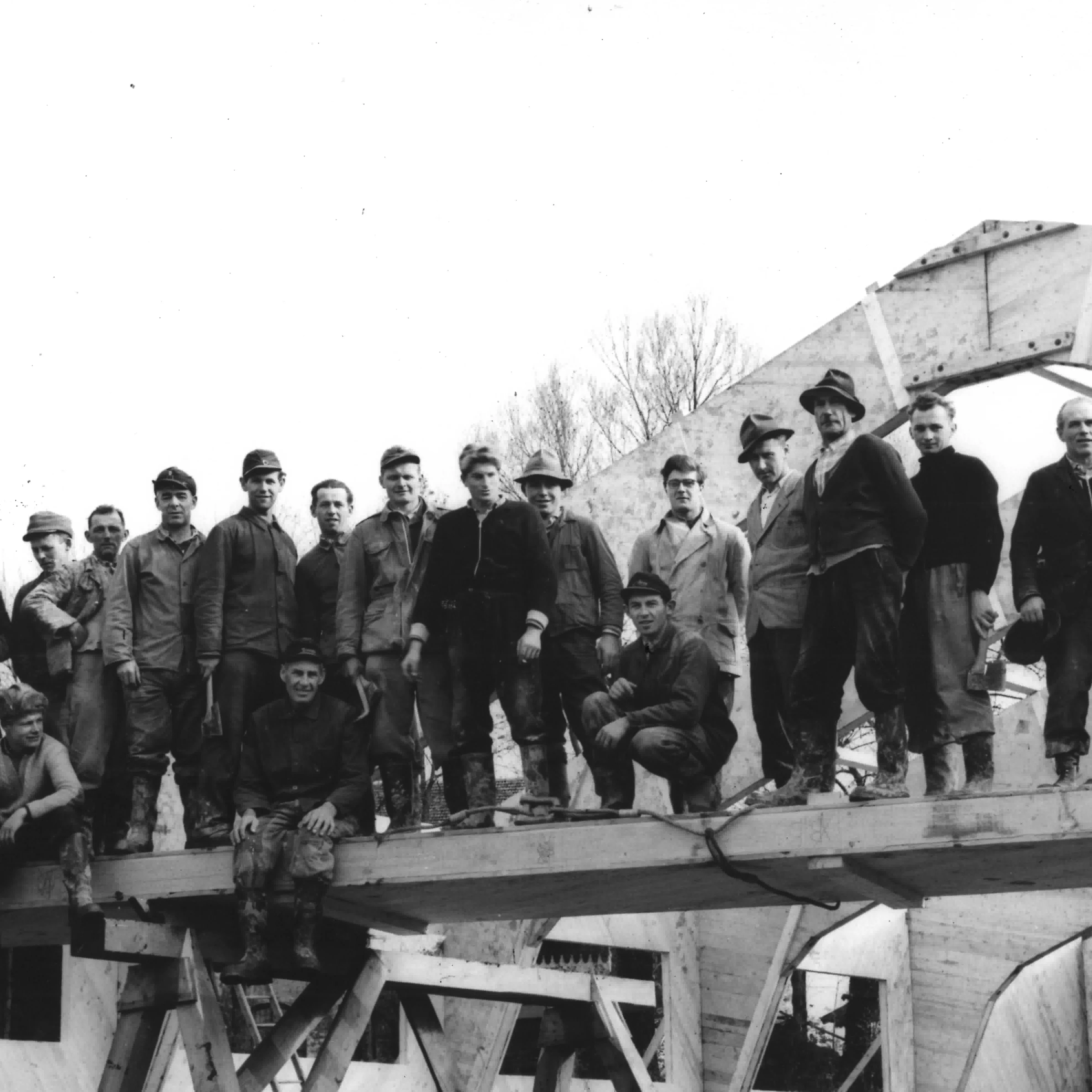 Schwarz-Weiß-Foto einer Gruppe von 15 Bauarbeitern, die stolz vor ihrem Bauprojekt posieren. Einige der Männer tragen Kappen und Hüte, die Kleidung ist robust und typisch für die harte Arbeit. Die Männer stehen oder hocken auf Holzbalken und im Hintergrund sind weitere Bauteile zu sehen.