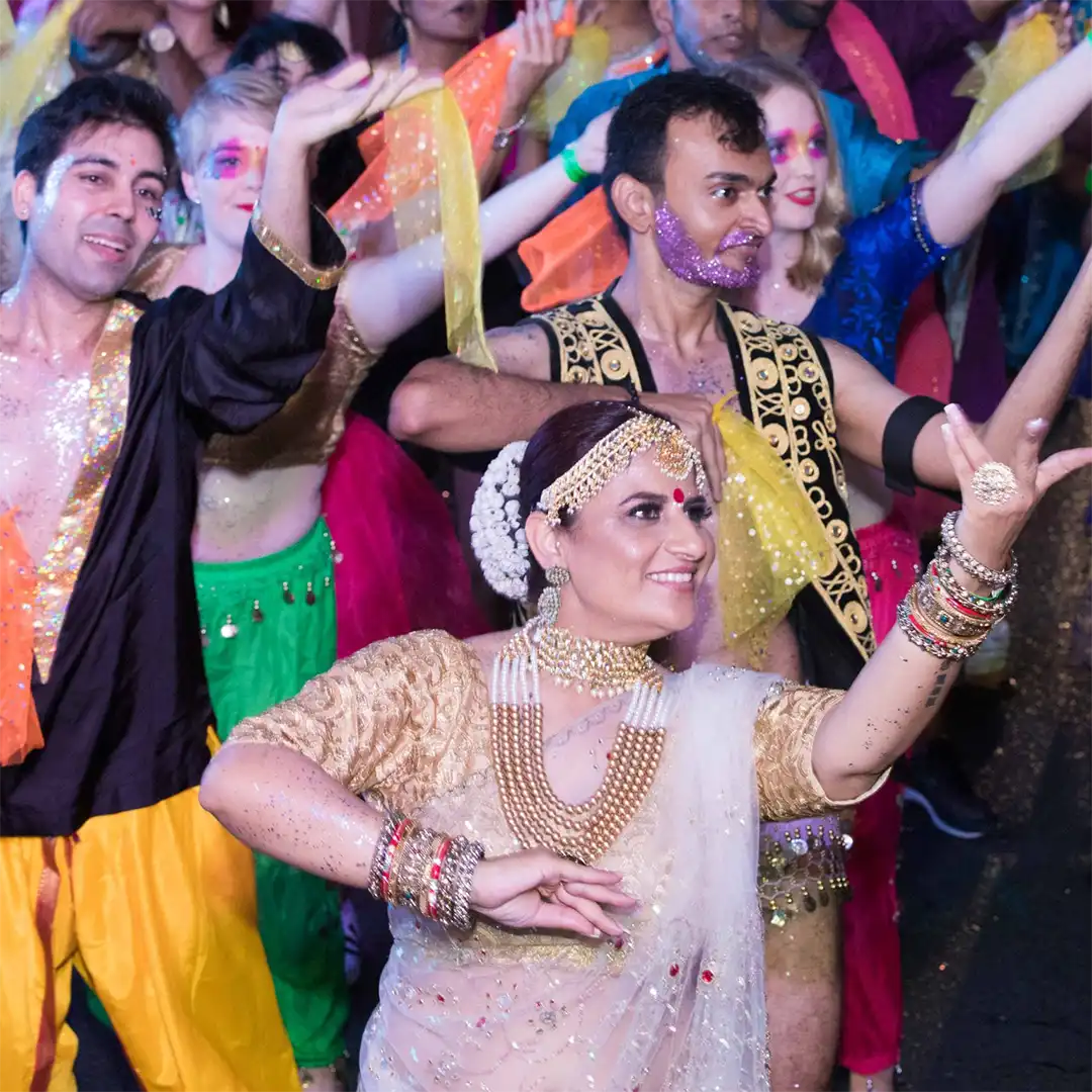 A group of people in colourful, glittering costumes dance joyfully, with a woman in traditional Indian attire and jewellery leading at the front. Bright scarves and festive make-up add to the lively atmosphere.