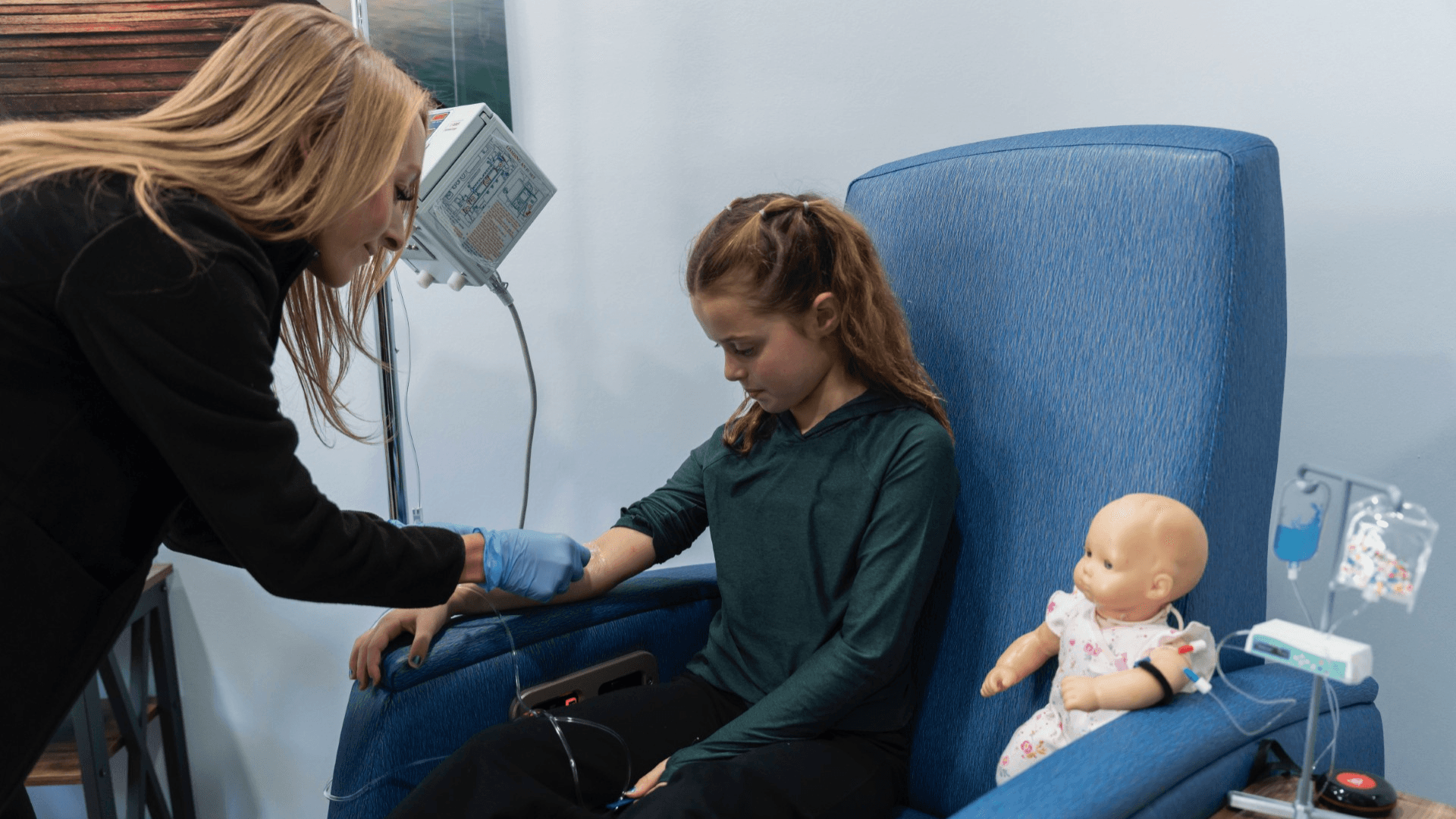 Healthcare professional placing an IV line in a young girl’s arm while she sits in a hospital chair, with a doll beside her also set up with a mock IV