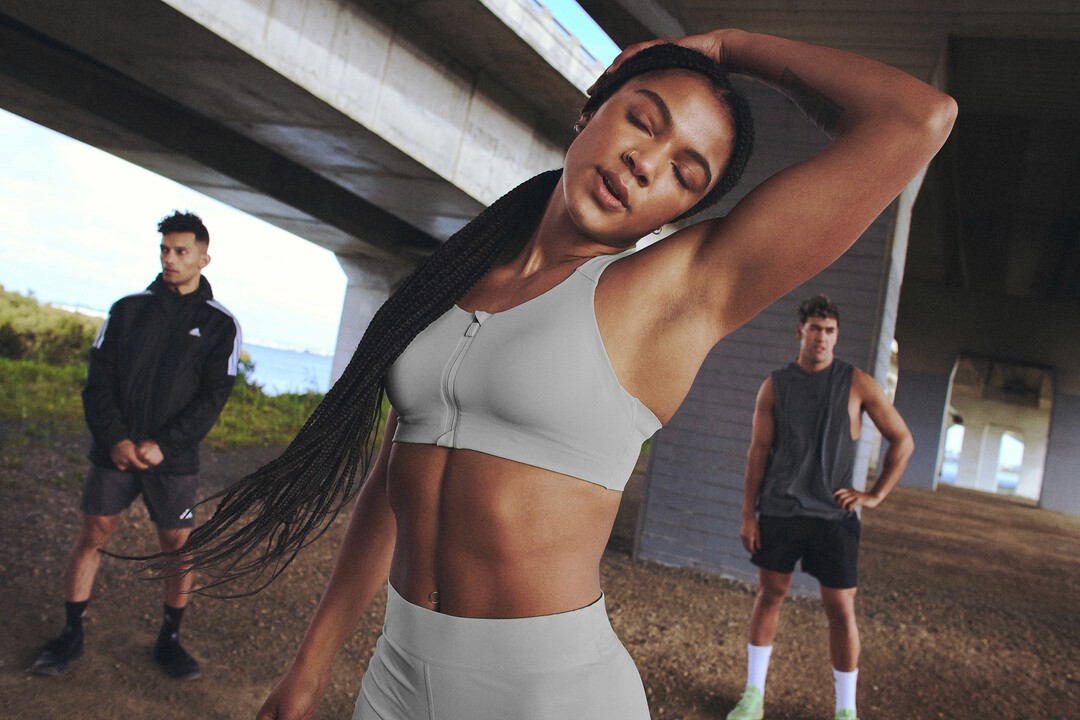 Frau in Sport-Kleidung greift sich mit der linken Hand über den Kopf und dehnt sich den Nacken. Im Hintergrund stehen zwei Männer in Sportkleidung..